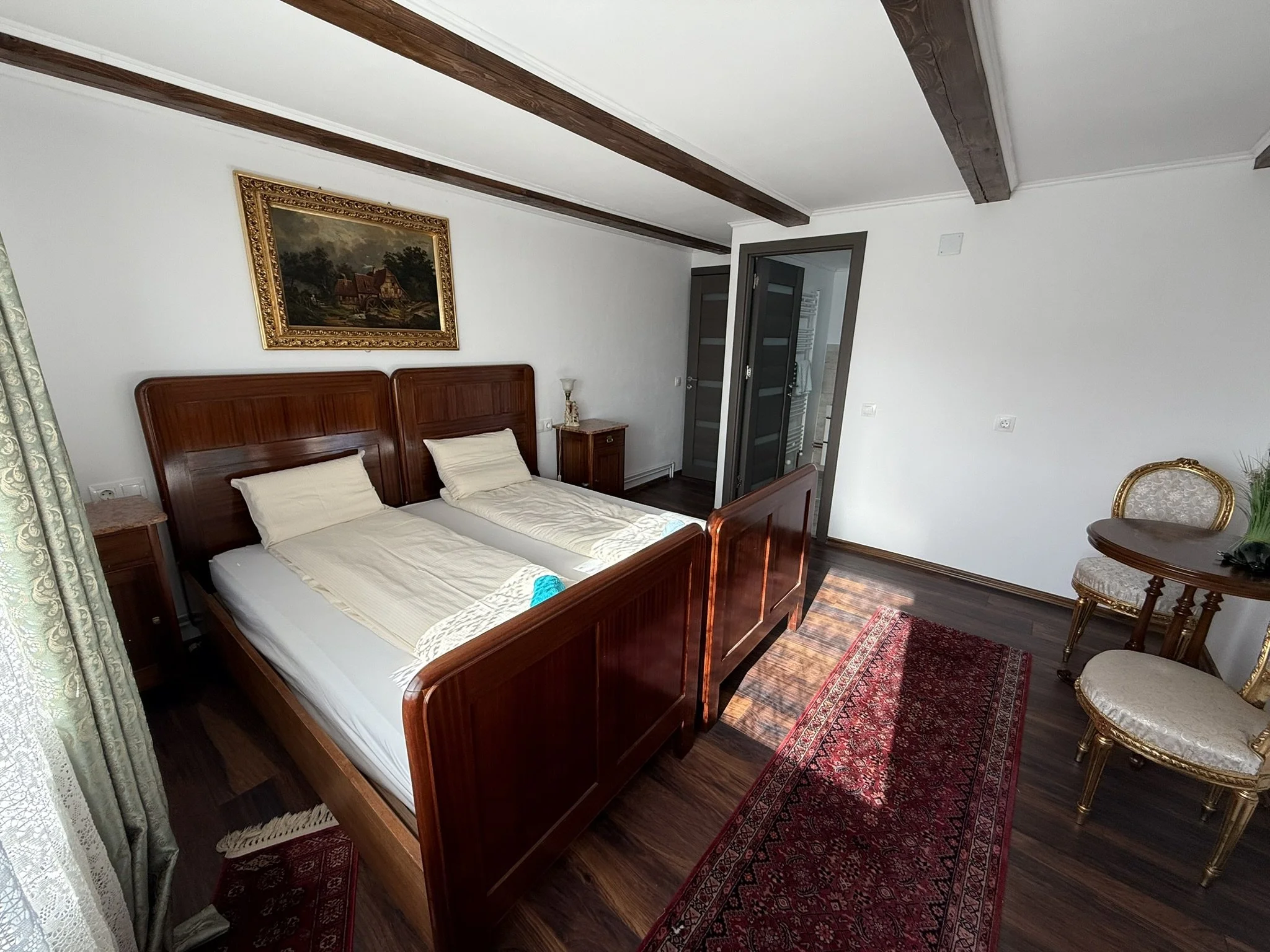 A bedroom with two twin beds with wooden headboards, a bedside table with a lamp, a painting above the beds, and a small sitting area with two ornate chairs and a small round table. The room has dark wood flooring and carpet, with a partially open do