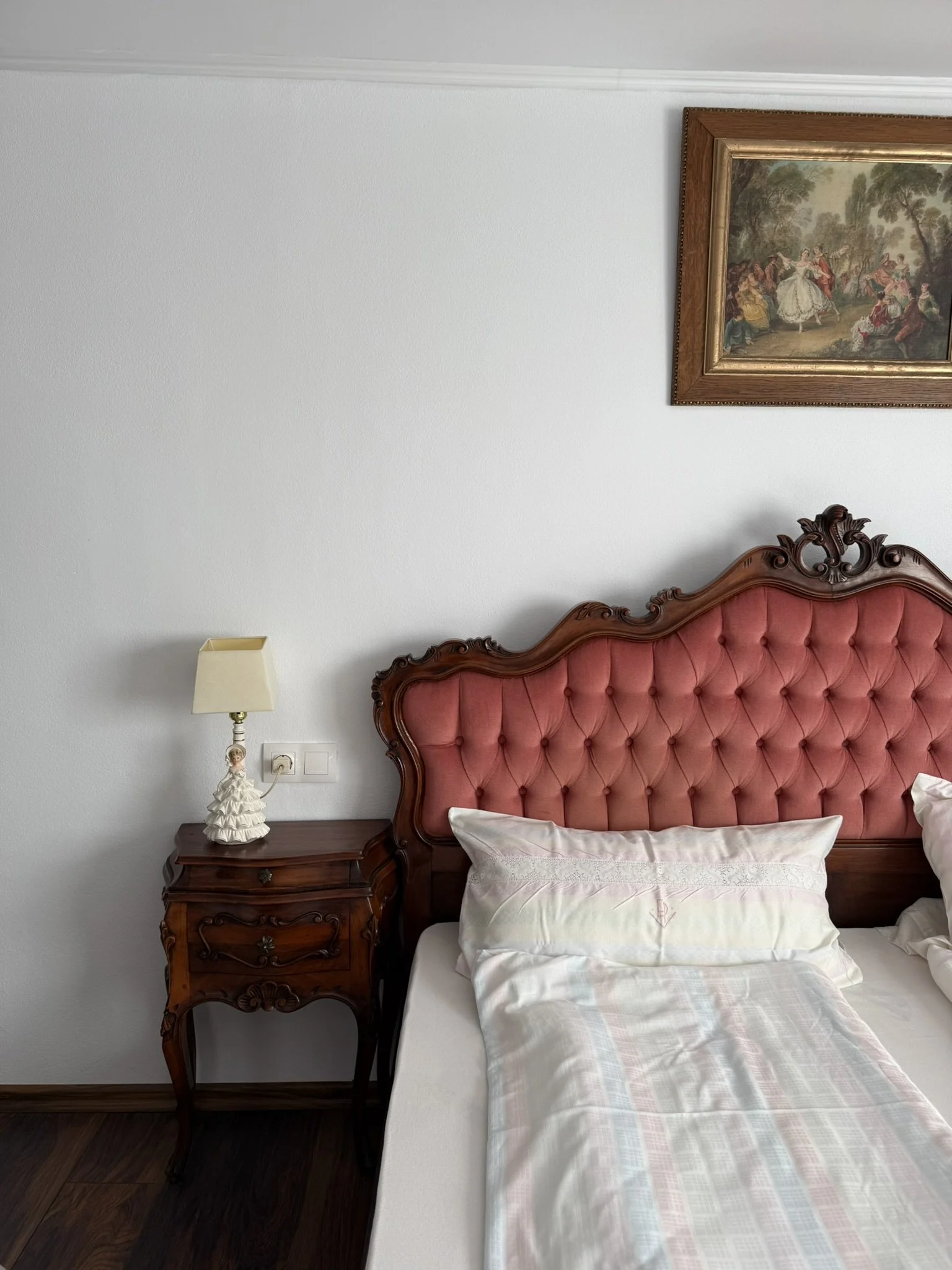 A bedroom featuring a vintage wooden headboard with pink upholstery, a peacefully made bed with white pillows and a light-colored sheet, a small wooden nightstand with ornate carvings, a table lamp with a cream shade and a ceramic base shaped like a 