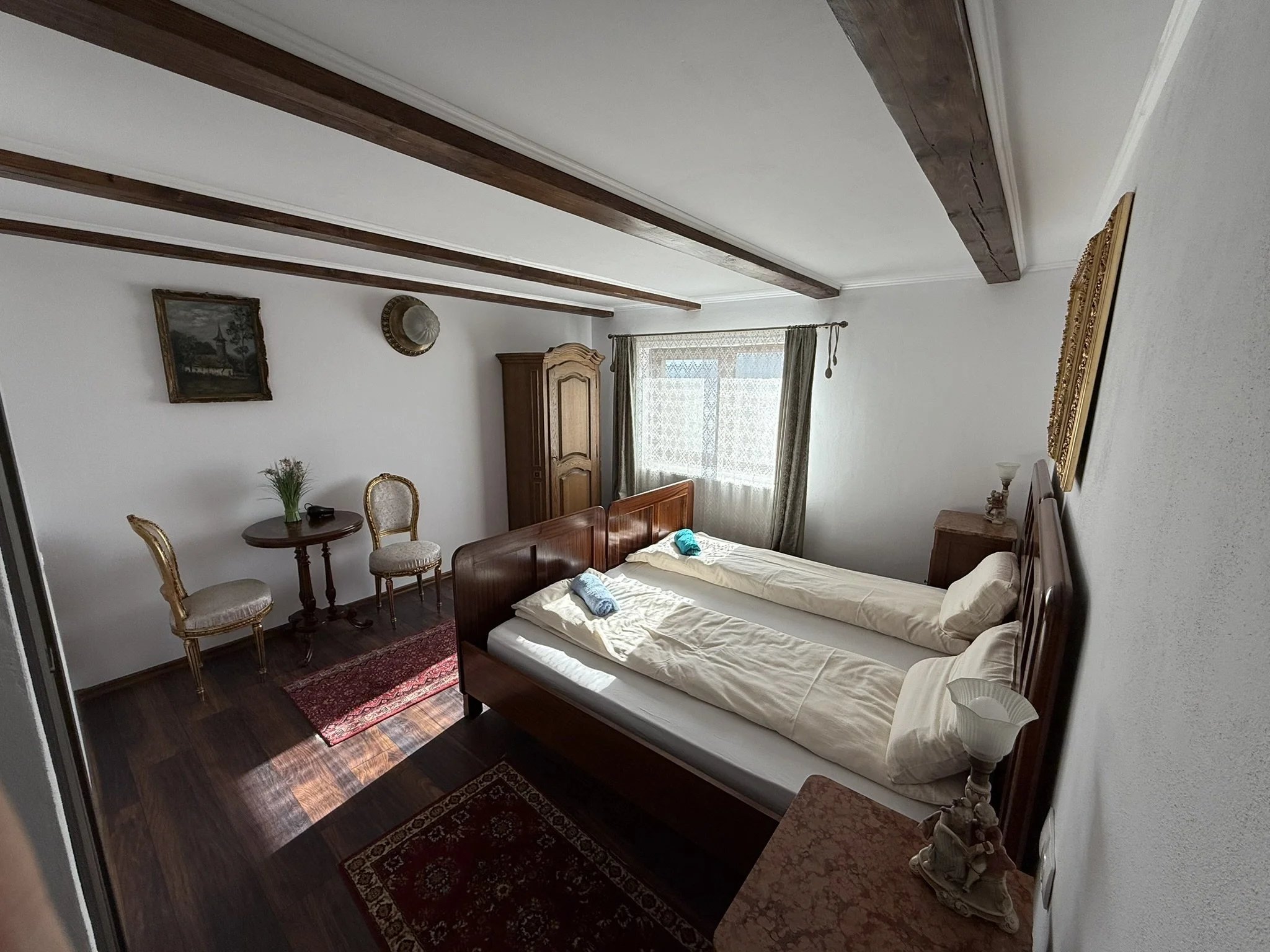 A bedroom with two beds, a window with curtains, a small round table with two chairs, a wardrobe, and wall decorations.