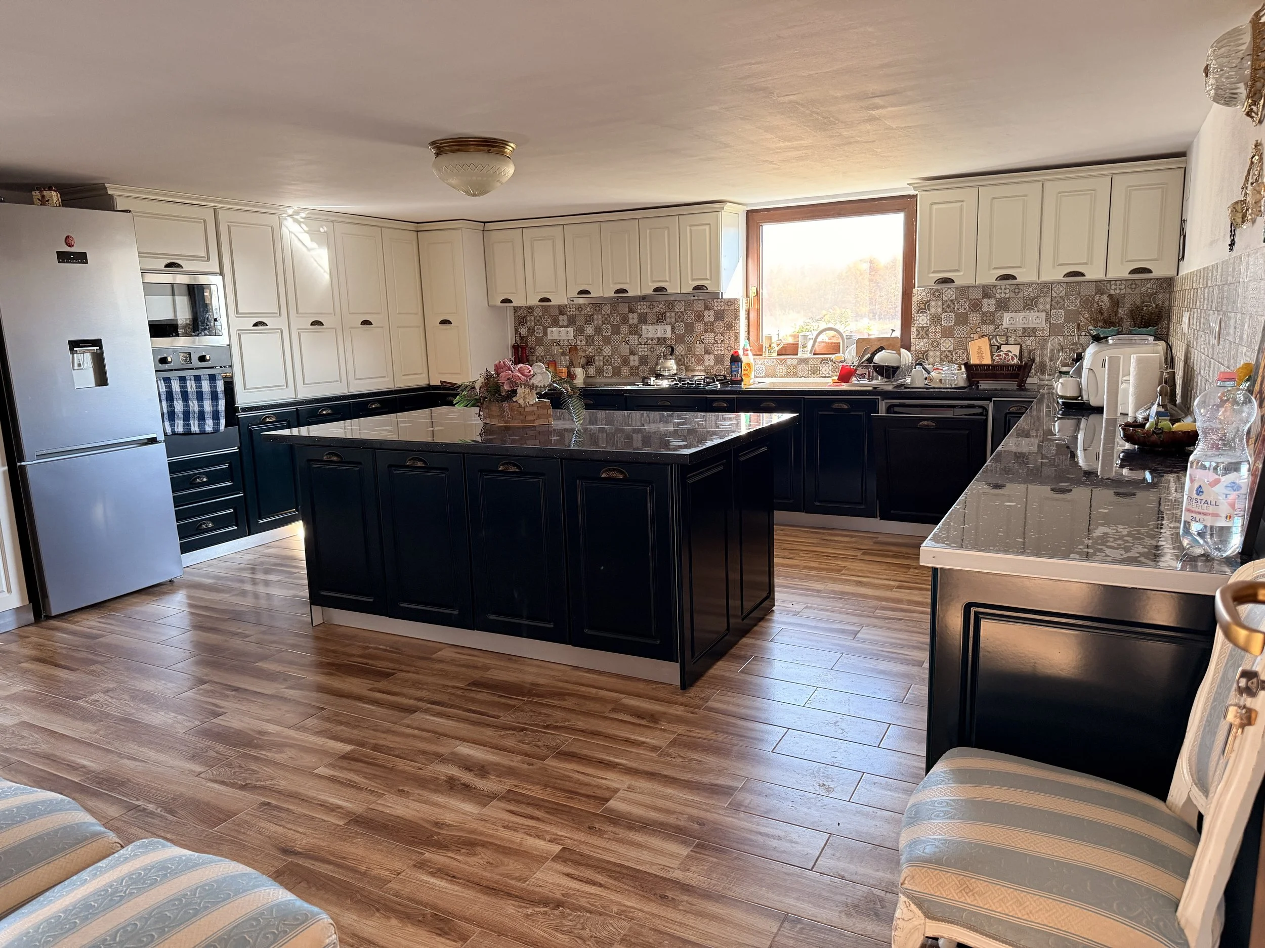 A spacious kitchen with white upper cabinets and black lower cabinets, a central black island, a stainless steel refrigerator, a window above the sink, and various small appliances on the countertop.