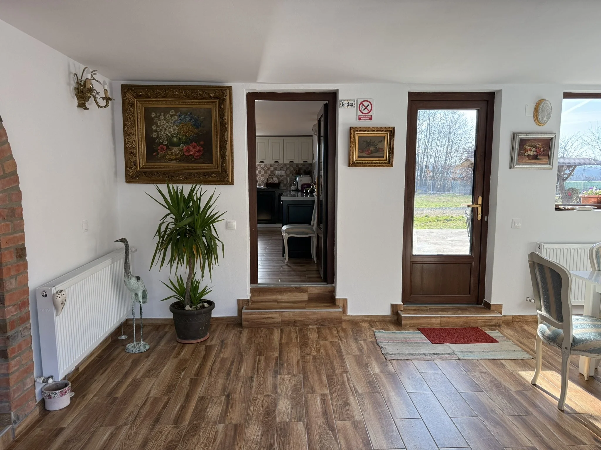 Interior of a home with wooden flooring, white walls, and a view through a glass-paneled door to an outdoor area with grass and trees. There are framed paintings on the wall, a potted plant, a sculpture, and a white dining chair.