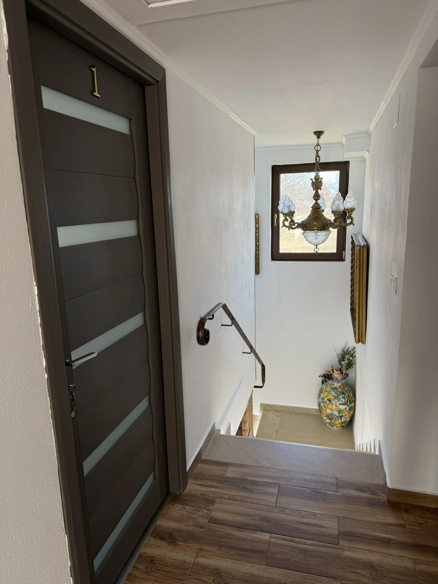 View of a staircase hallway with a closed gray front door numbered 1, a hanging chandelier light fixture, a window with a view outside, a decorative vase with flowers, and wooden flooring.