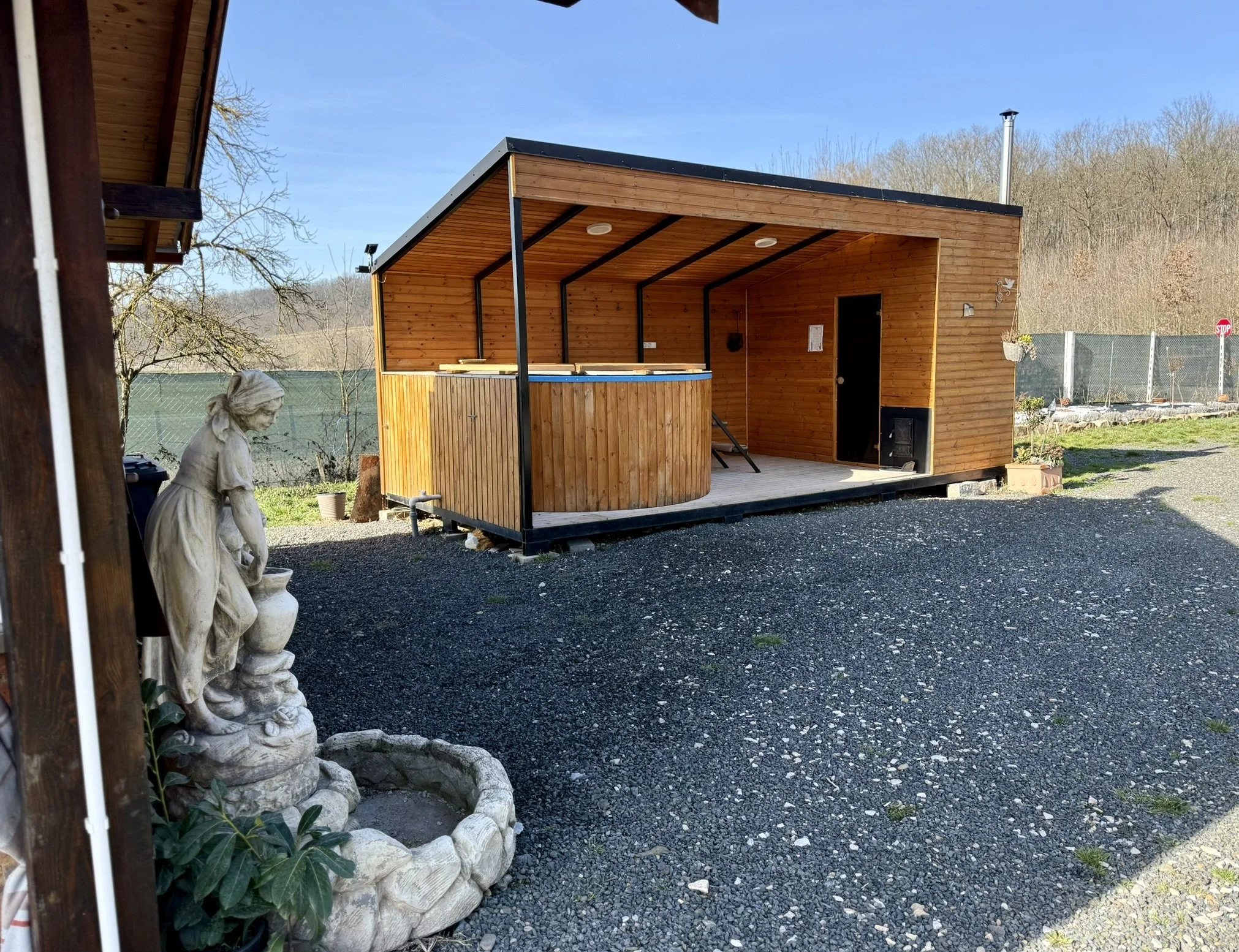 An outdoor wooden structure with a hot tub, situated on a gravel area, with a decorative statue of a woman holding a water jug in the foreground, and trees in the background.
