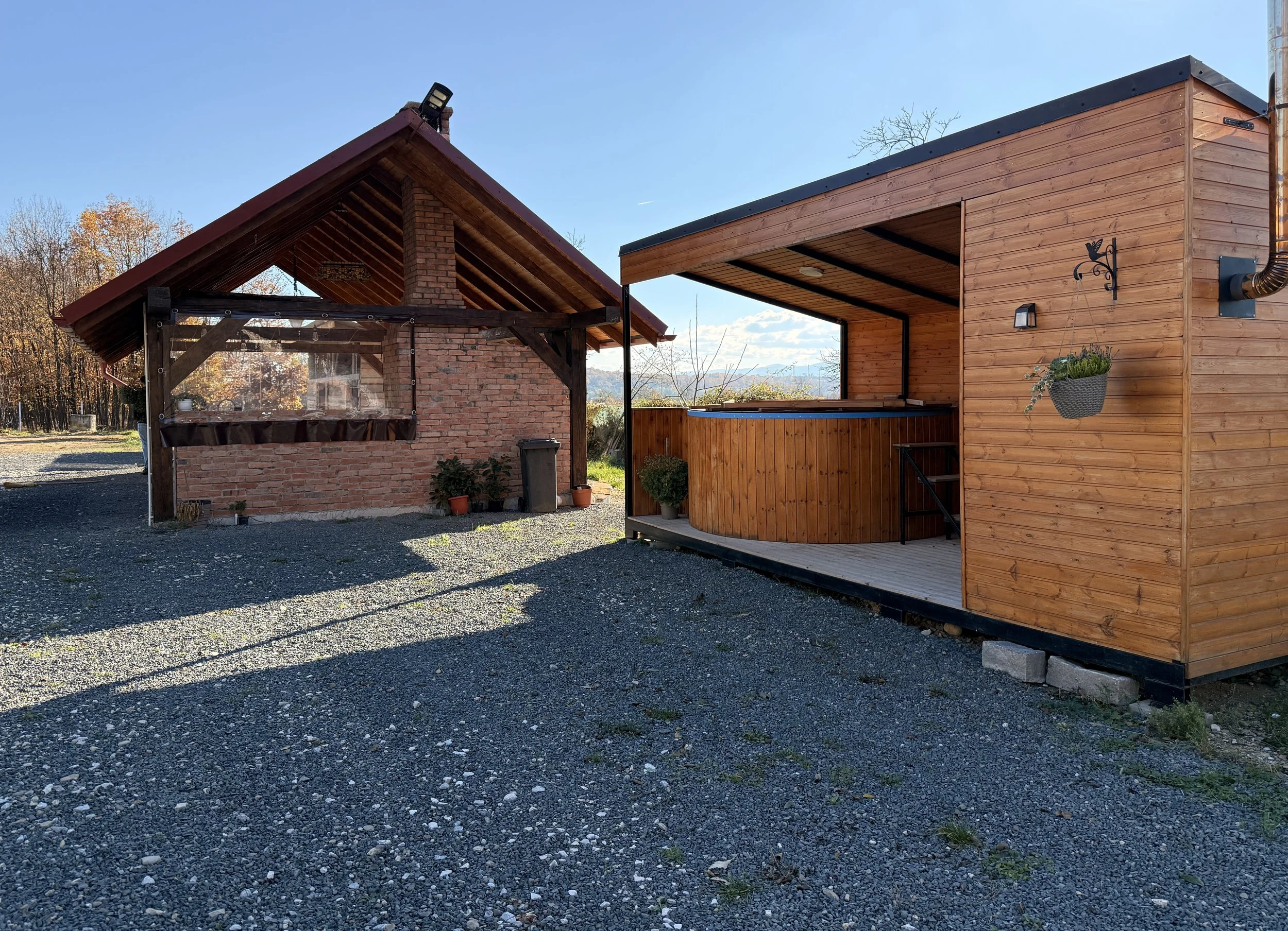 A photo of two adjacent outdoor structures, one with a brick wall and wooden roof, bbq, and the other with wooden paneling and a small covered deck, jacuzzi and sauna under a clear blue sky with trees in the background.
