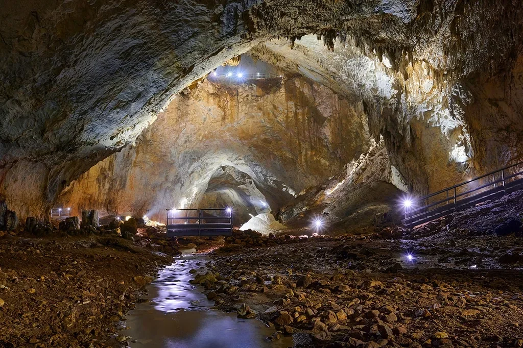 Inside a large, illuminated cave with rocky walls, guided walkway, and shallow stream, lit with artificial lighting.