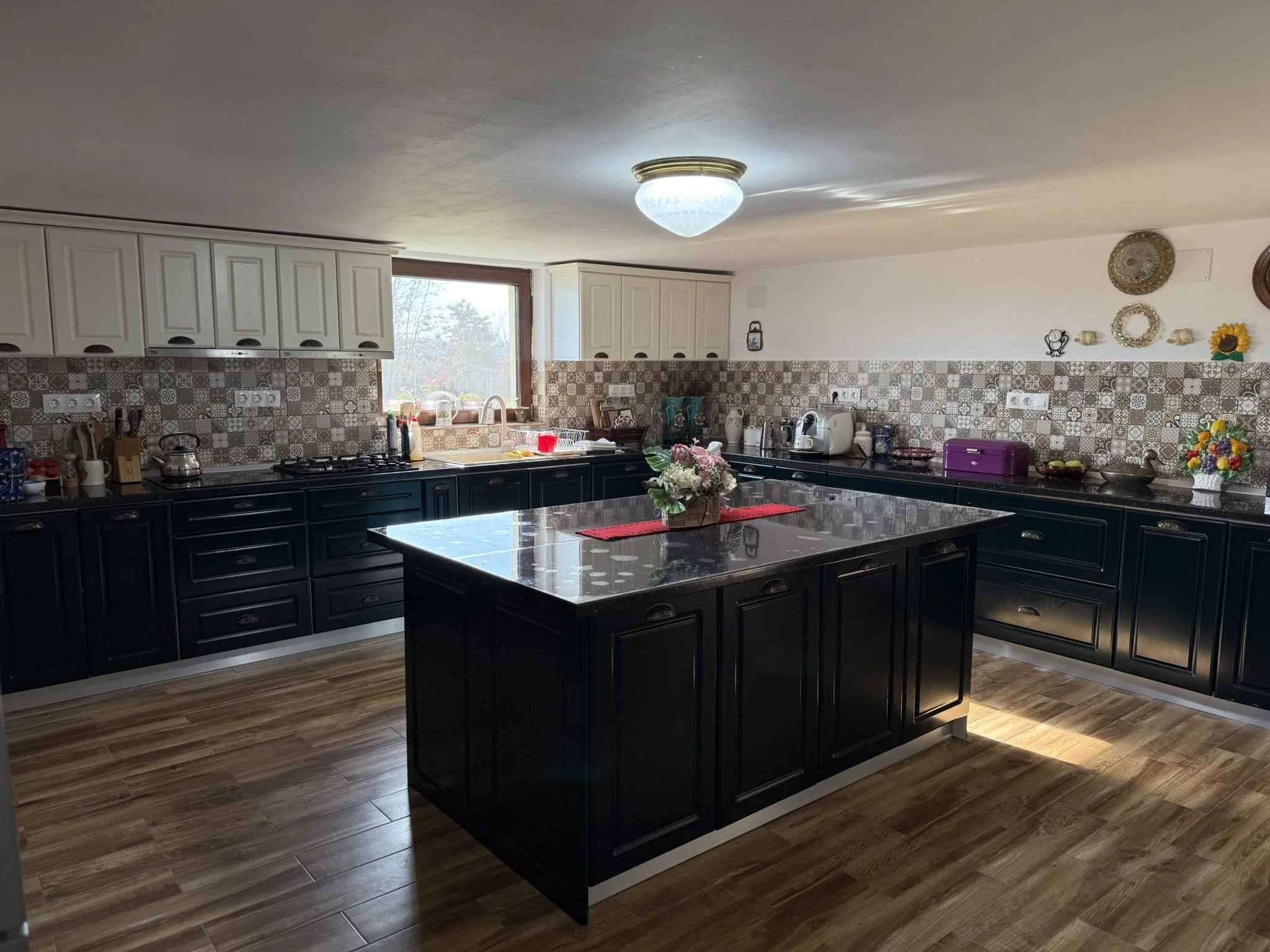 Kitchen with black island and white upper cabinets, patterned backsplash, wooden flooring, window above sink, various appliances and decorative items on countertops.