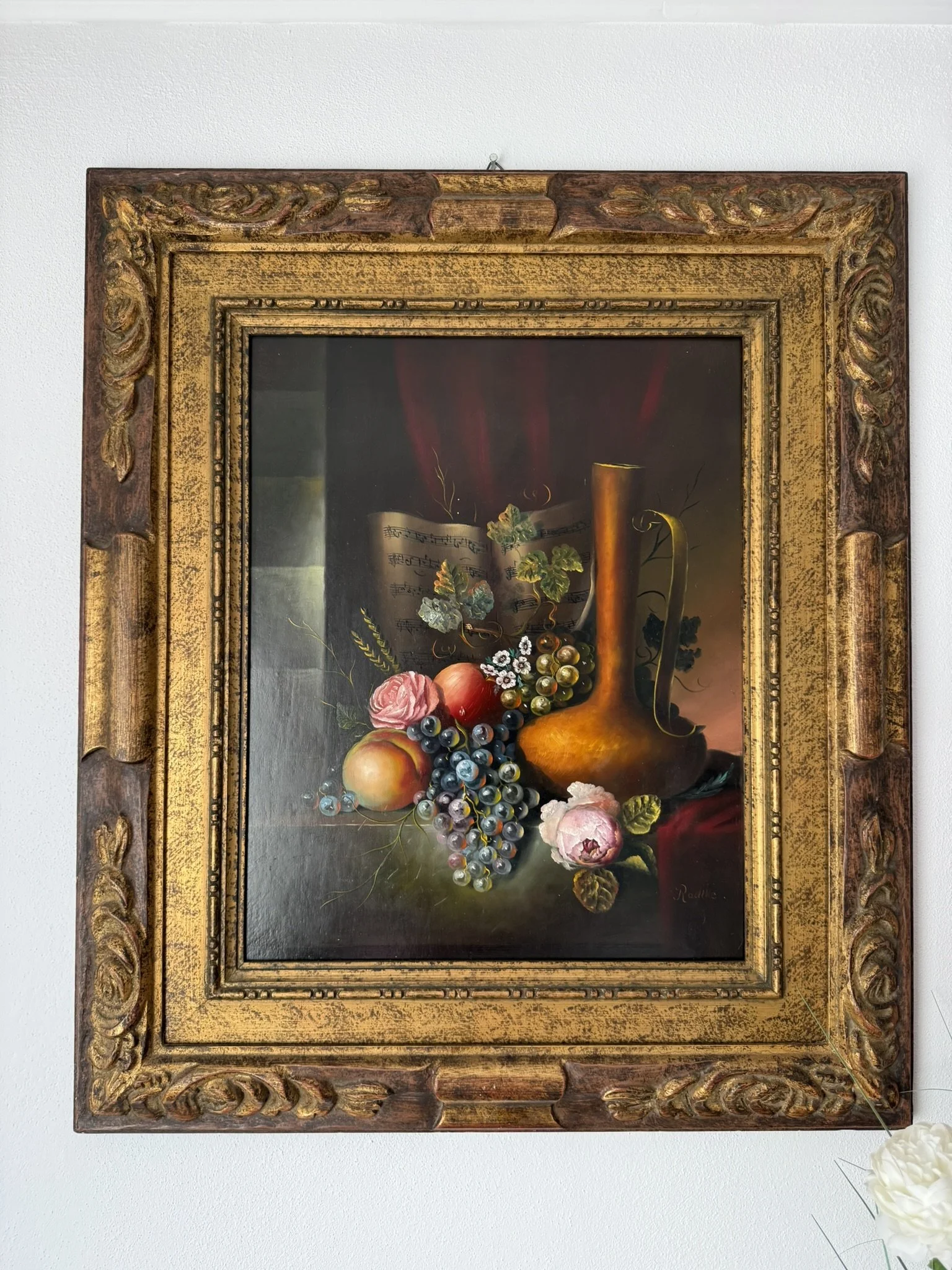 A still life painting of fruit, flowers, and a golden pitcher with sheet music in the background, framed in an ornate gold and wood frame.
