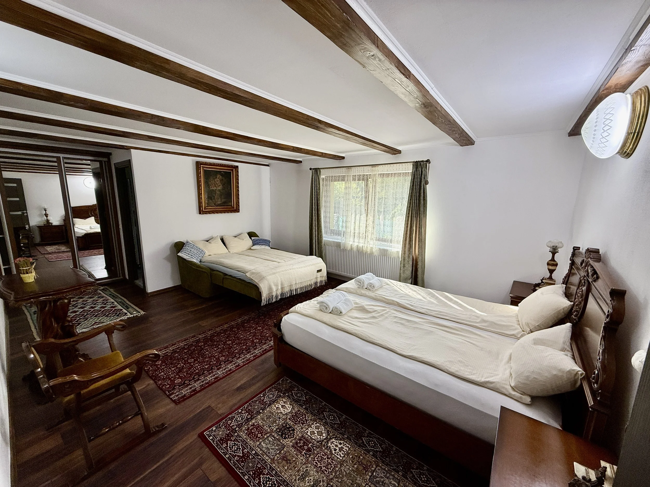 Hotel bedroom with two beds, one larger and one smaller, with rolled towels on both beds, wooden furniture, patterned rugs, a window with green curtains, and a partial view into an adjoining room with a bedroom in the background.