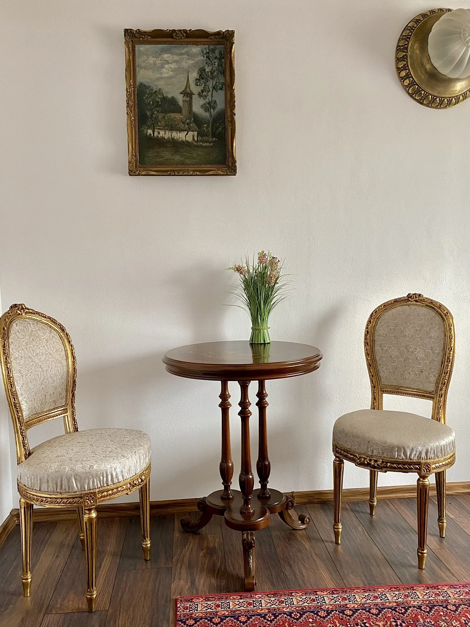 A small round wooden table with a vase of pink flowers, flanked by two upholstered chairs with gold frames, in a room with hardwood floors, a white wall, a framed landscape painting, and a decorative wall sconce.