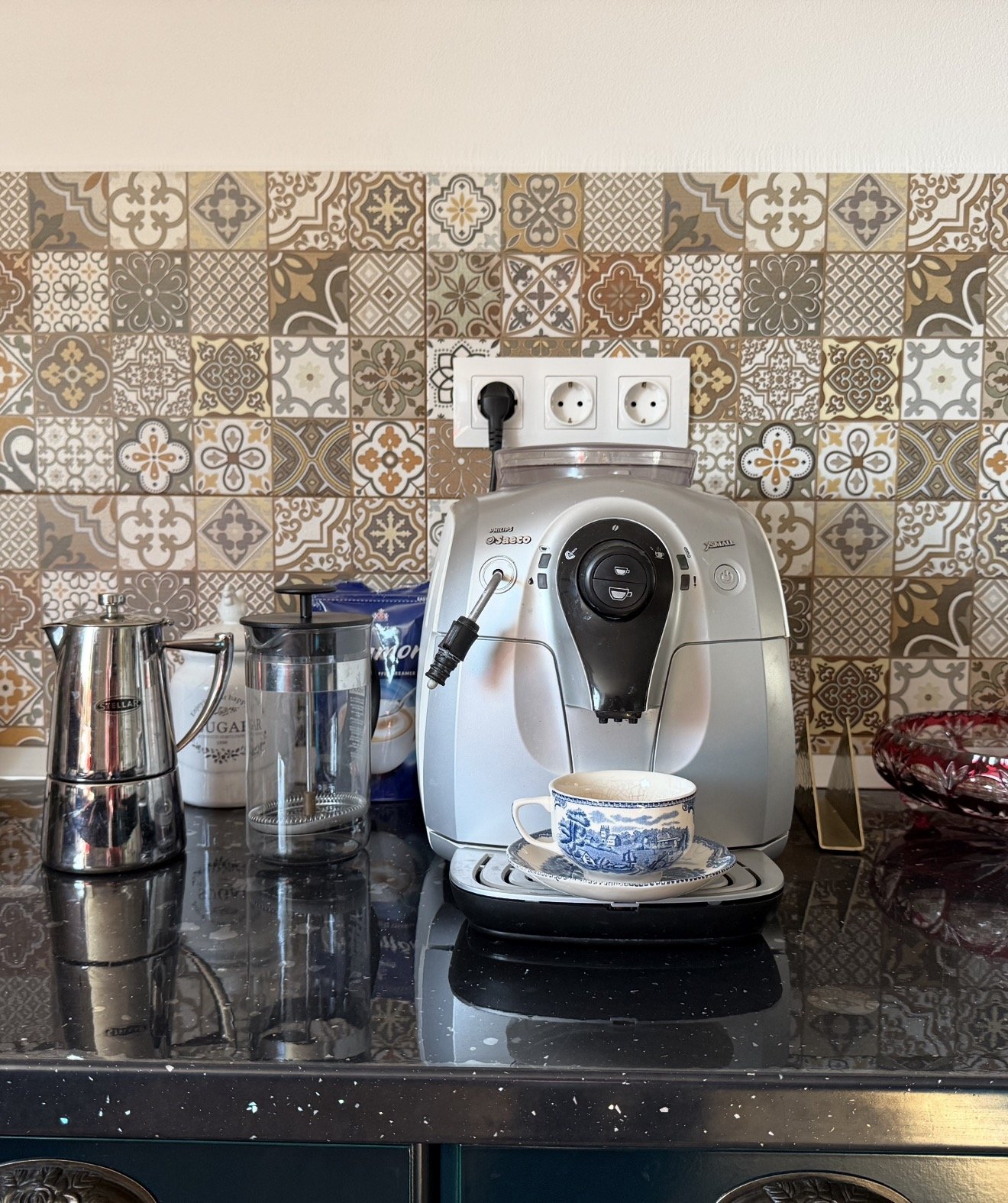 Coffee machine with a cup, placed on a black marble kitchen counter, alongside a sugar jar, a grinder, and a bag of coffee, with a colorful patterned tile backsplash in the background.