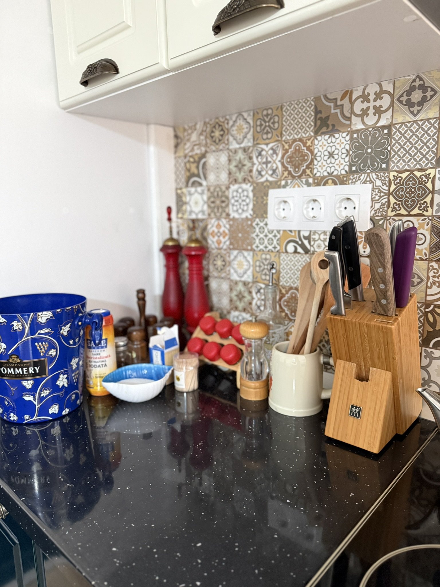 A kitchen countertop with various utensils, spices, and containers, including a knife block with knives, salt and pepper shakers, and a decorative jar.