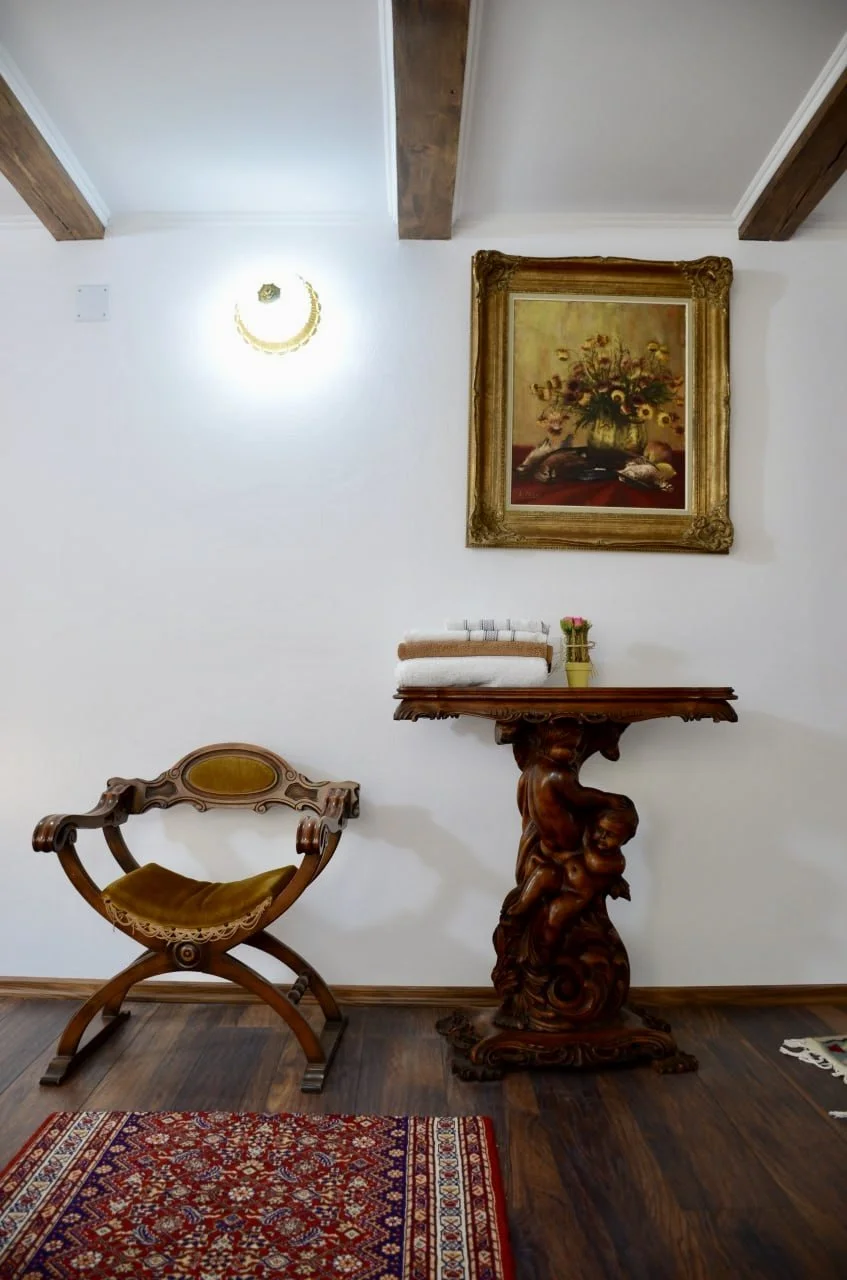 Interior of a room with a wooden chair, decorative wooden table with a carved pedestal base, a framed floral painting on the wall, a stack of towels, a small vase with flowers, and a decorative rug on dark hardwood floor.