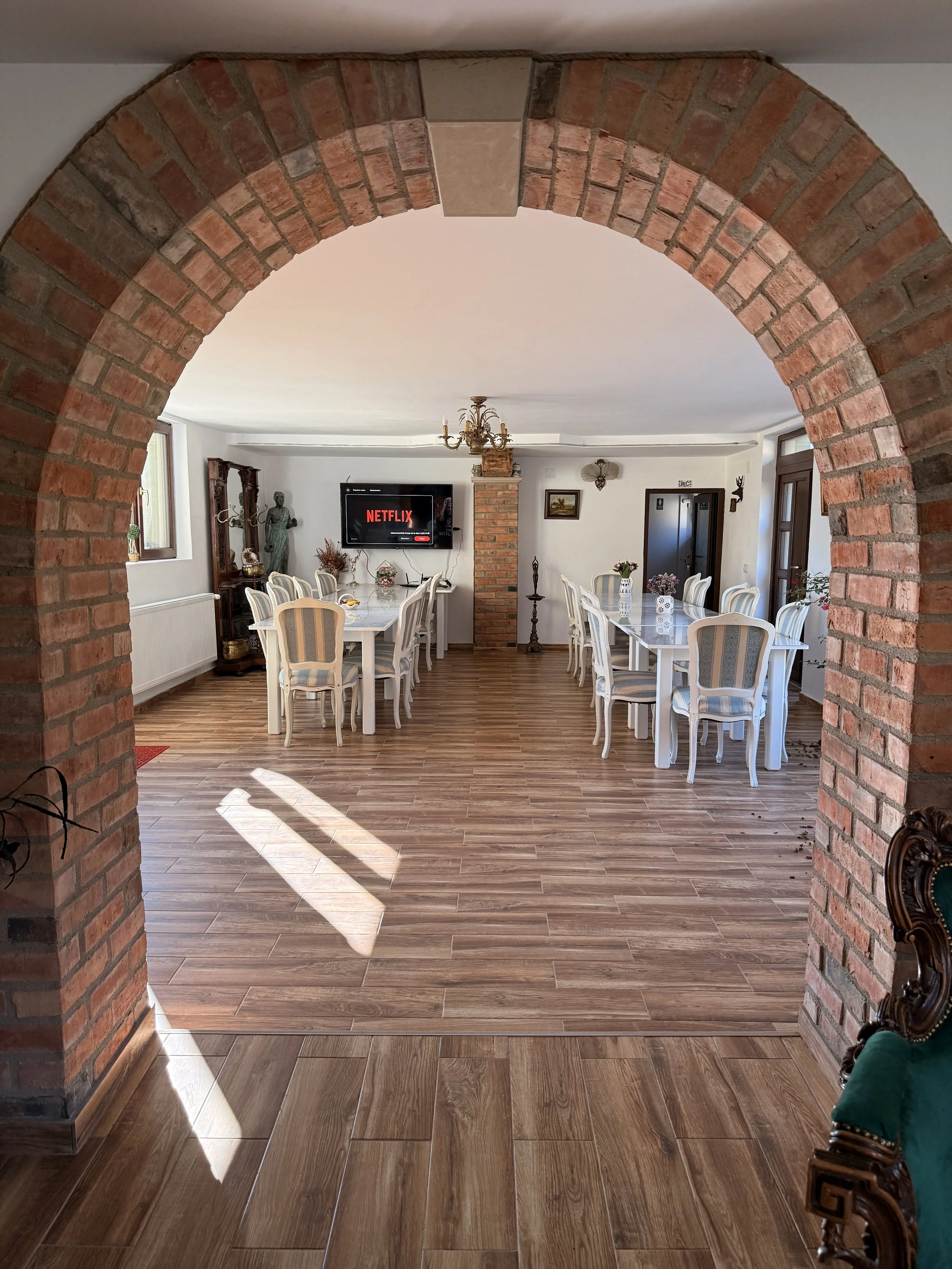 A spacious dining room viewed through a brick archway, with two large white dining tables surrounded by cream and blue upholstered chairs, hardwood floors, a flat-screen TV on the wall displaying the Netflix logo, and decorative items including a sta