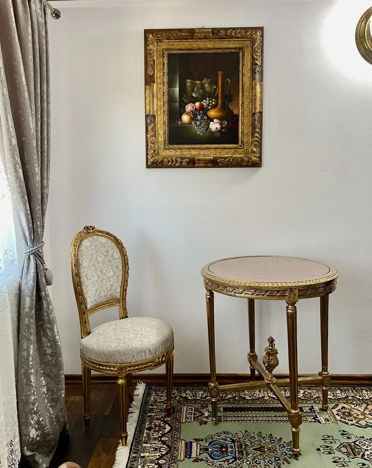 An ornate room corner with a gold framed painting of a still life of fruit and a vase, a gold and cream upholstered chair, a matching small round table, patterned curtains, and a decorative rug.
