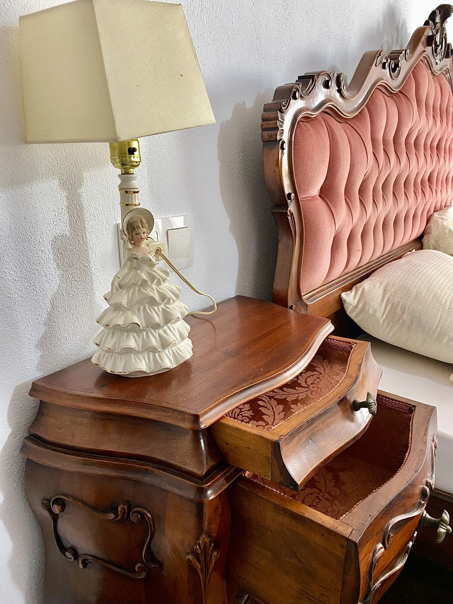A wooden nightstand with a vintage ceramic lamp and a pink, tufted headboard with carved detailing in a bedroom.