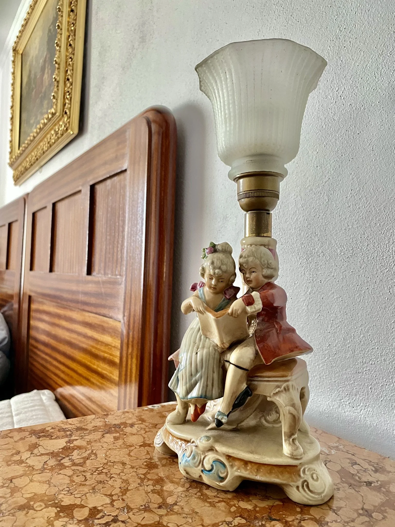 A vintage ceramic table lamp with two children reading a book at the base, placed on a brown marble surface in a room with a wooden headboard and a framed picture on the wall.