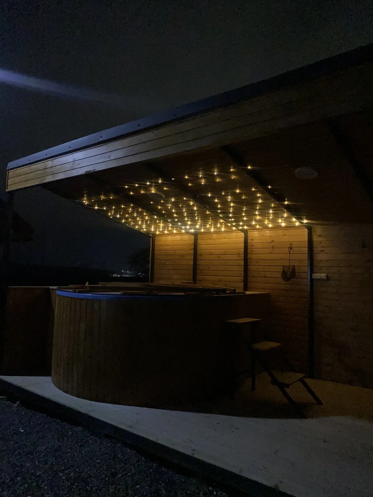 Night view of a wooden hot tub with a roof, decorated with string lights, on a patio area.
