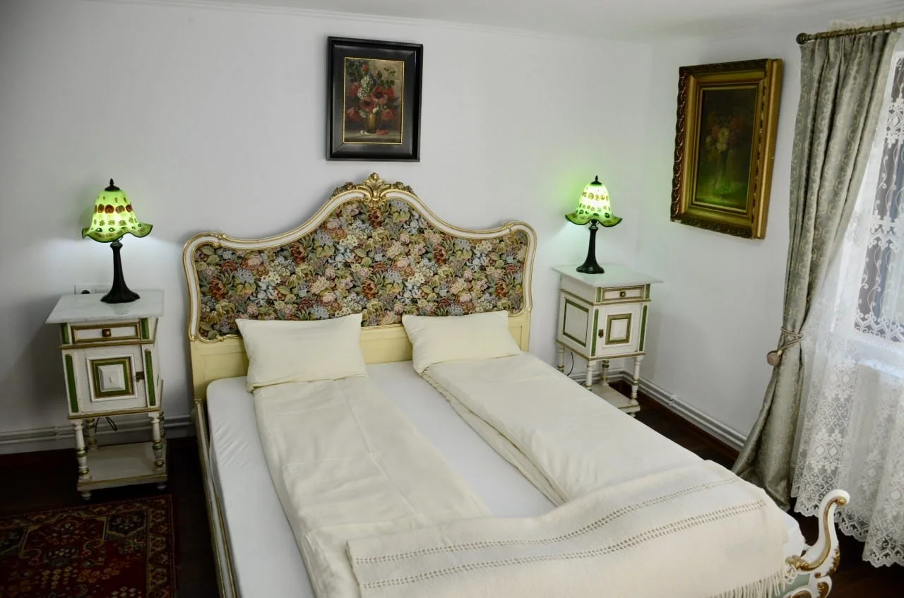A vintage-style bedroom with a double bed featuring a floral upholstered headboard, two cream pillows, and cream sheets. There are two matching white nightstands with green and gold accents on either side of the bed, each holding a green table lamp w