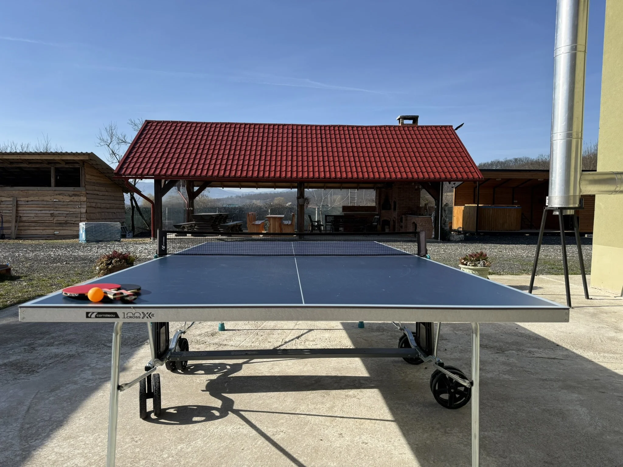 Outdoor table tennis setup with a black table, paddles, and an orange ball, under a red-roofed shelter with chairs, on a bright day with a gravel yard and wooden structures in the background.