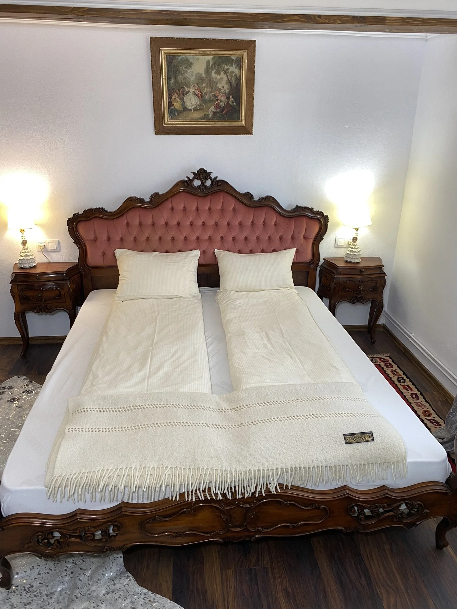 Vintage wooden bed with pink upholstered headboard, cream sheets, and two pillows in a bedroom with two nightstands and lamps on each side, a framed painting above the bed, and hardwood flooring.