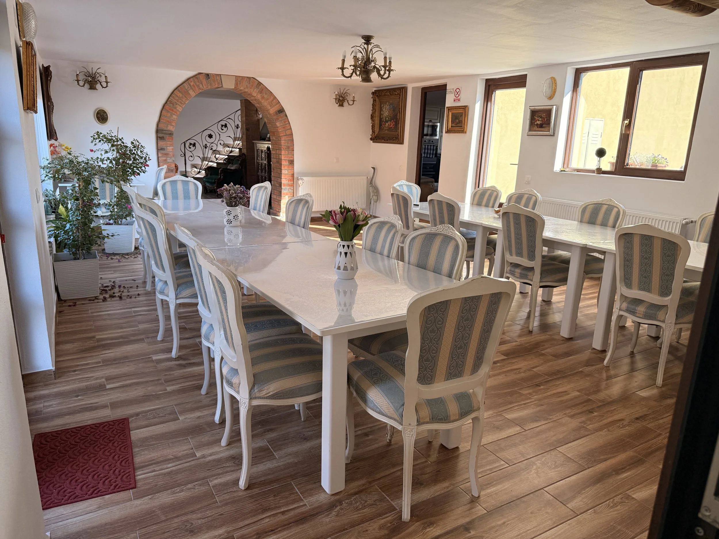 Dining room with long white marble table, 14 striped upholstered chairs, floral vases, and large windows letting in natural light.