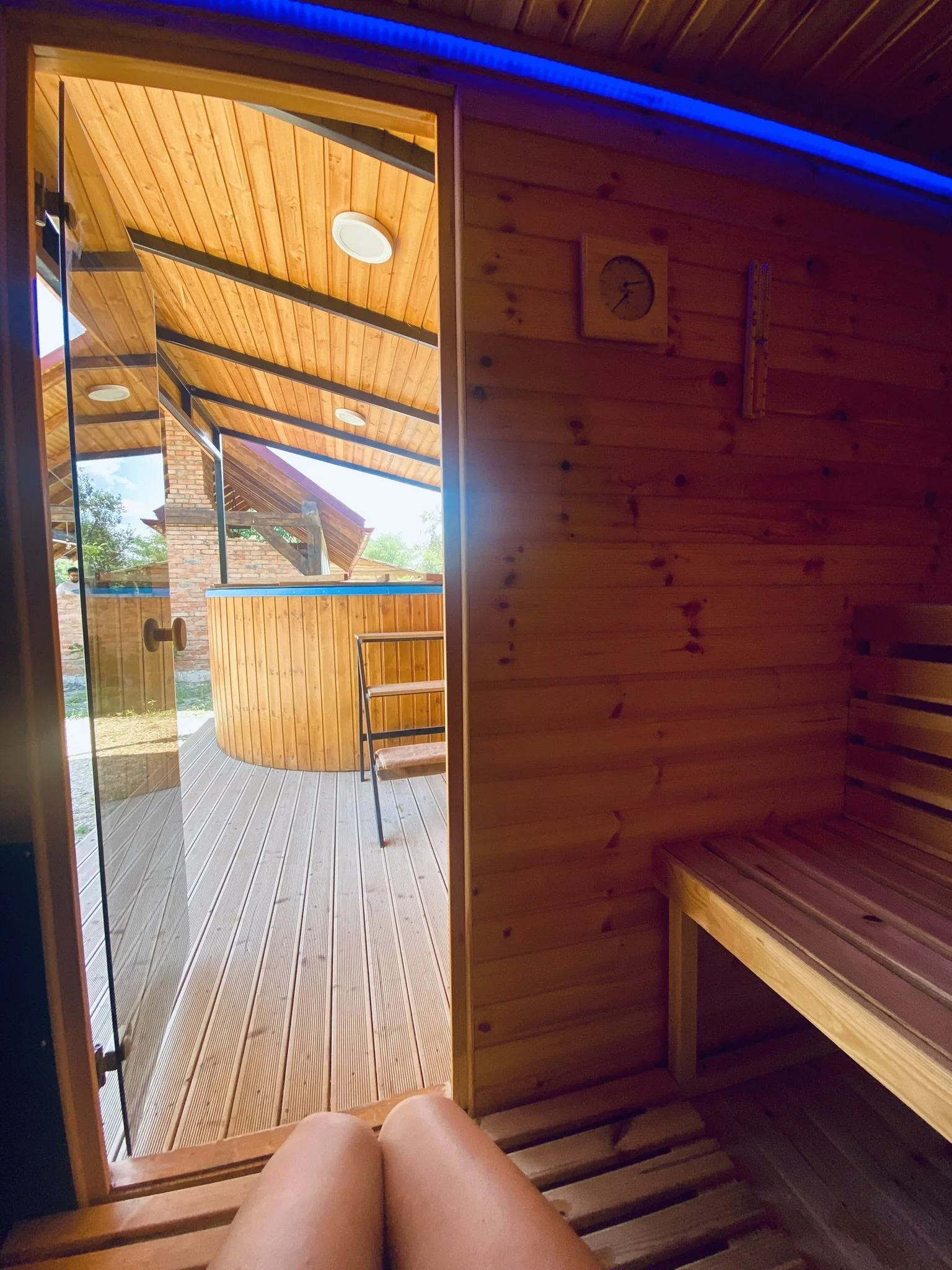 View from inside a wooden sauna looking out onto an outdoor deck with a hot tub, wood bench, and a chair, with a natural landscape and brick building in the background.