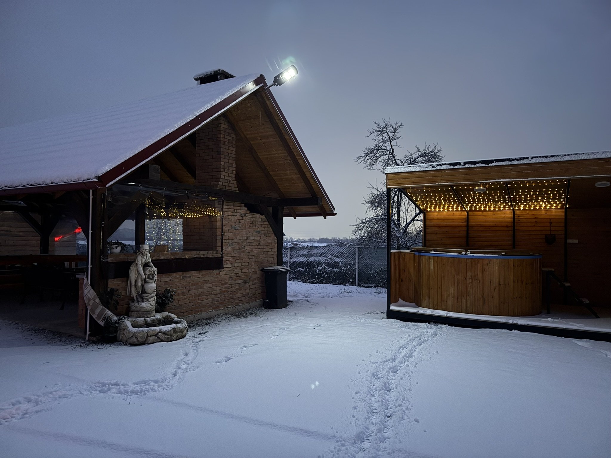 Snow-covered backyard with brick house and a hot tub under a wooden pergola, decorated with string lights, at dusk.