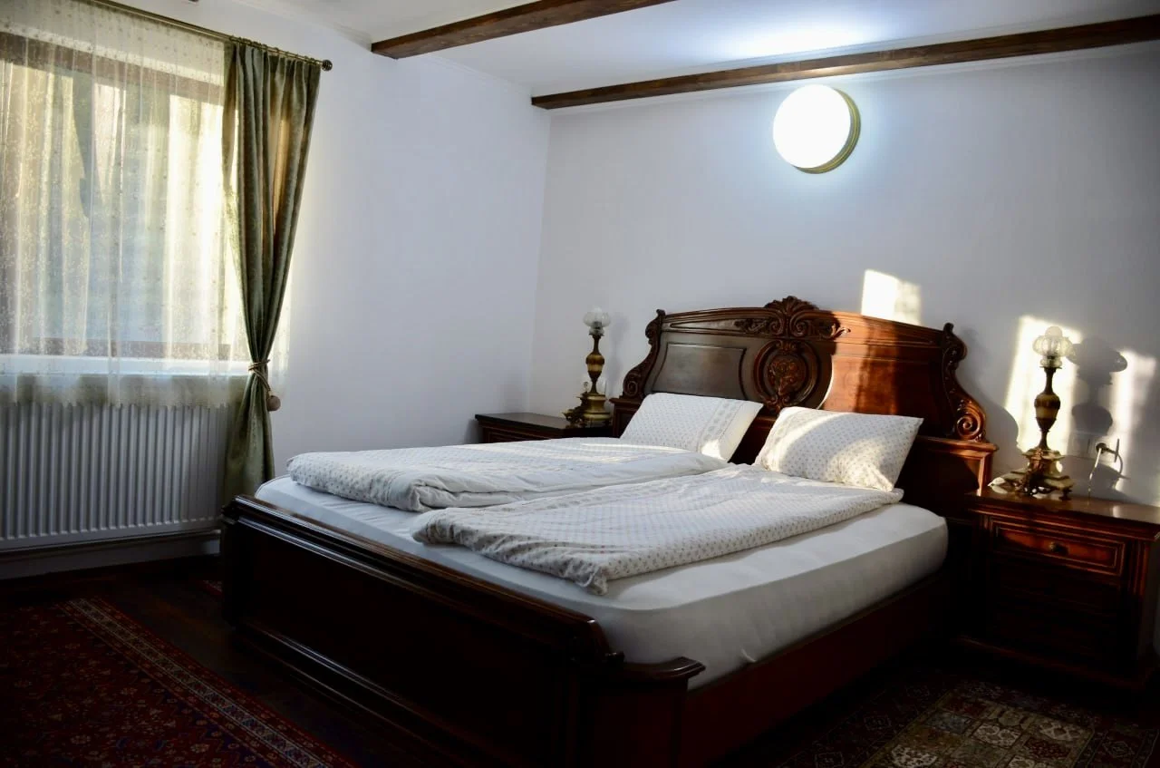 A cozy bedroom with a large wooden bed, two matching bedside tables with vintage lamps, a window with closed curtains, patterned rug, and sunflower-shaped wall light.