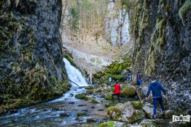 Group of people hiking through a rocky canyon with a small waterfall on the left and moss-covered rocks.