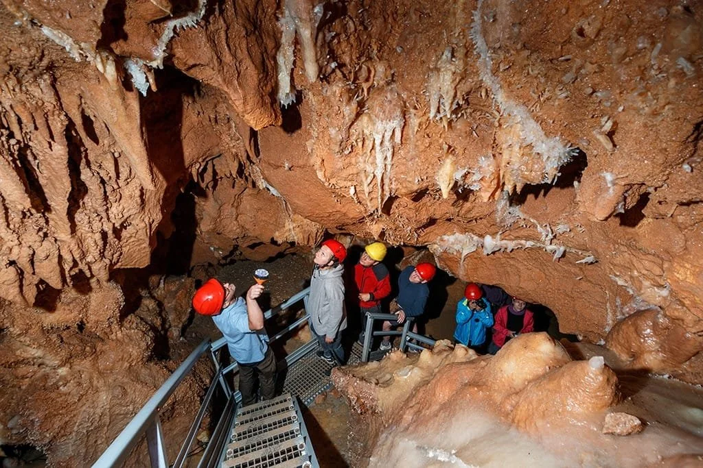 Group of people wearing helmets exploring a wet underground cave with reddish-brown rock formations.