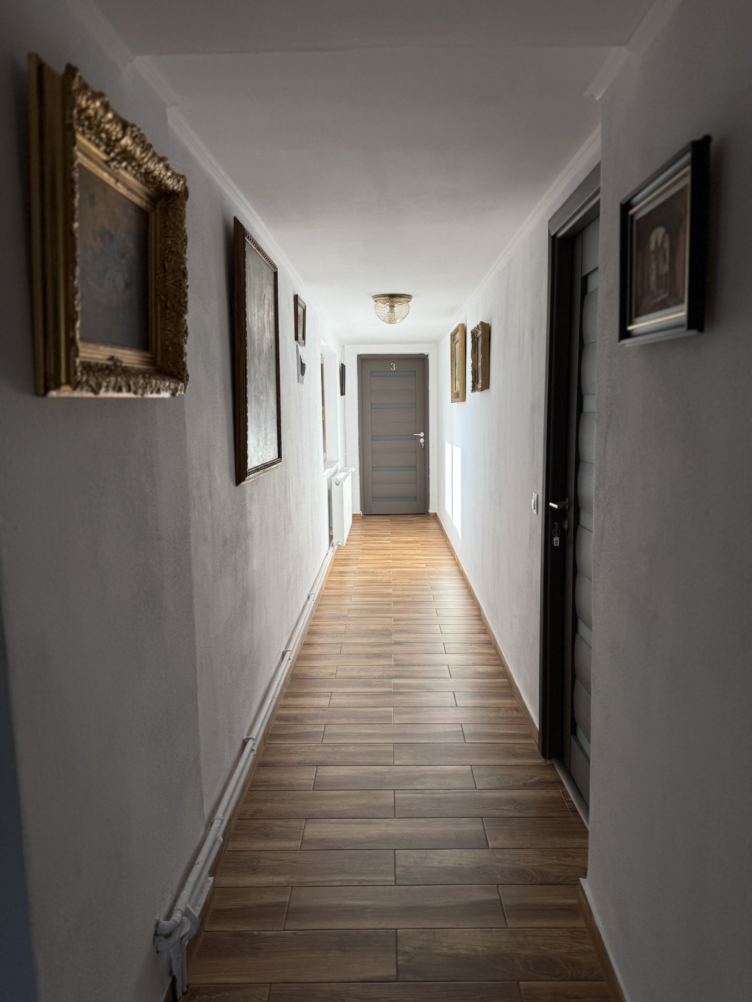 Long hallway with white walls, wooden floor, framed pictures on both sides, and a gray door at the end with the number 3.
