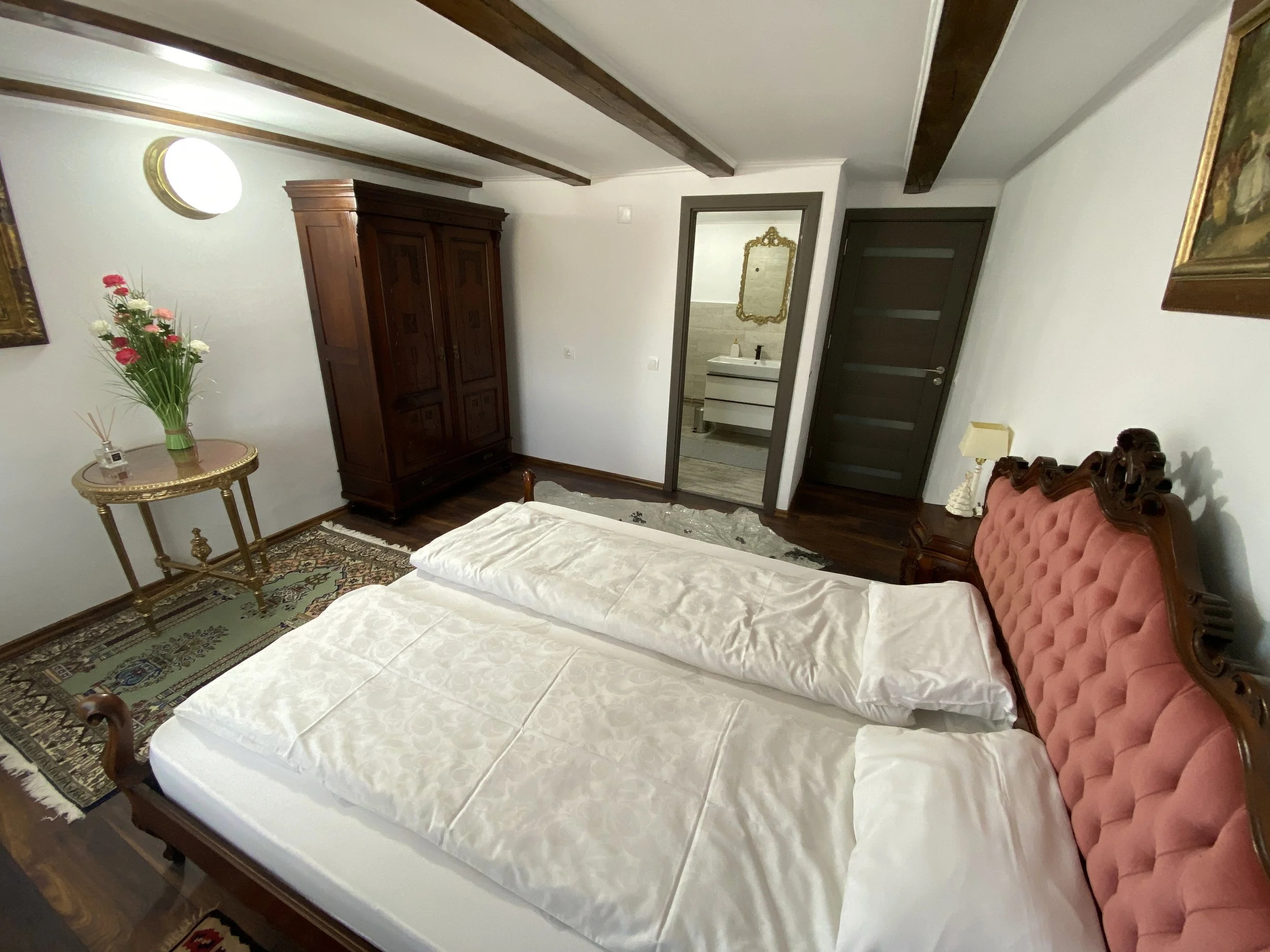 Hotel room with a large pink tufted headboard, wooden floor, and antique furniture including a wardrobe, side table with vase of flowers, and a small bedside lamp. The bed has white sheets and pillows, and there is a door leading to a bathroom with a