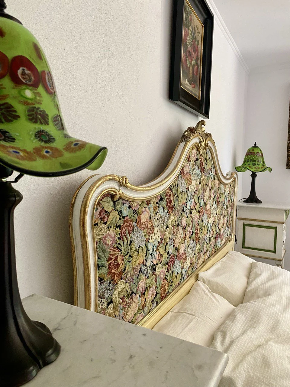 A bedroom with a floral upholstered headboard, two green and black lamps with floral designs, a framed painting of flowers on the wall, and a white nightstand with green trim.