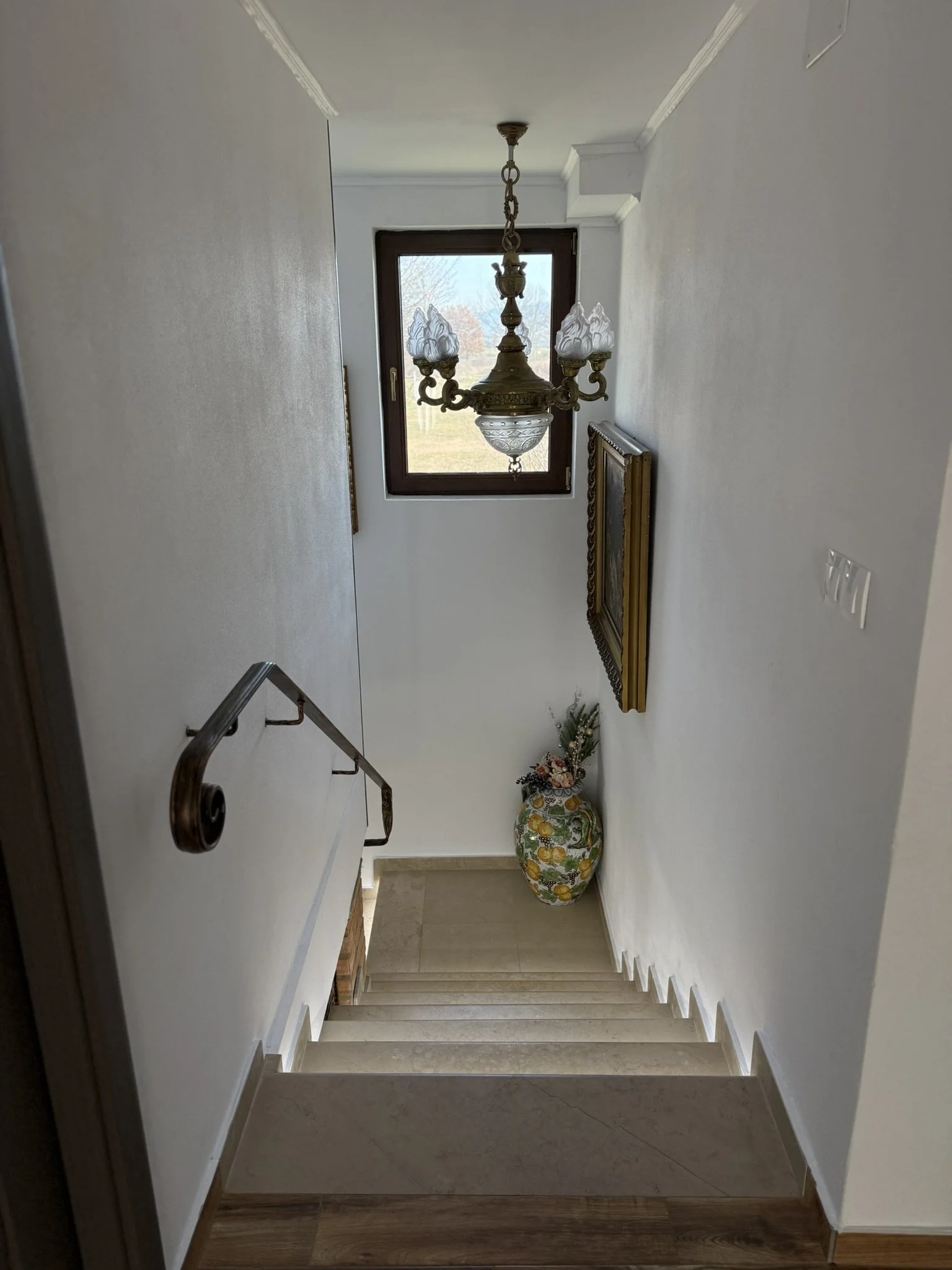 Interior view of a staircase in a house with a window at the landing, a chandelier hanging from the ceiling, a botanical vase with flowers on the floor, and framed artwork on the wall.