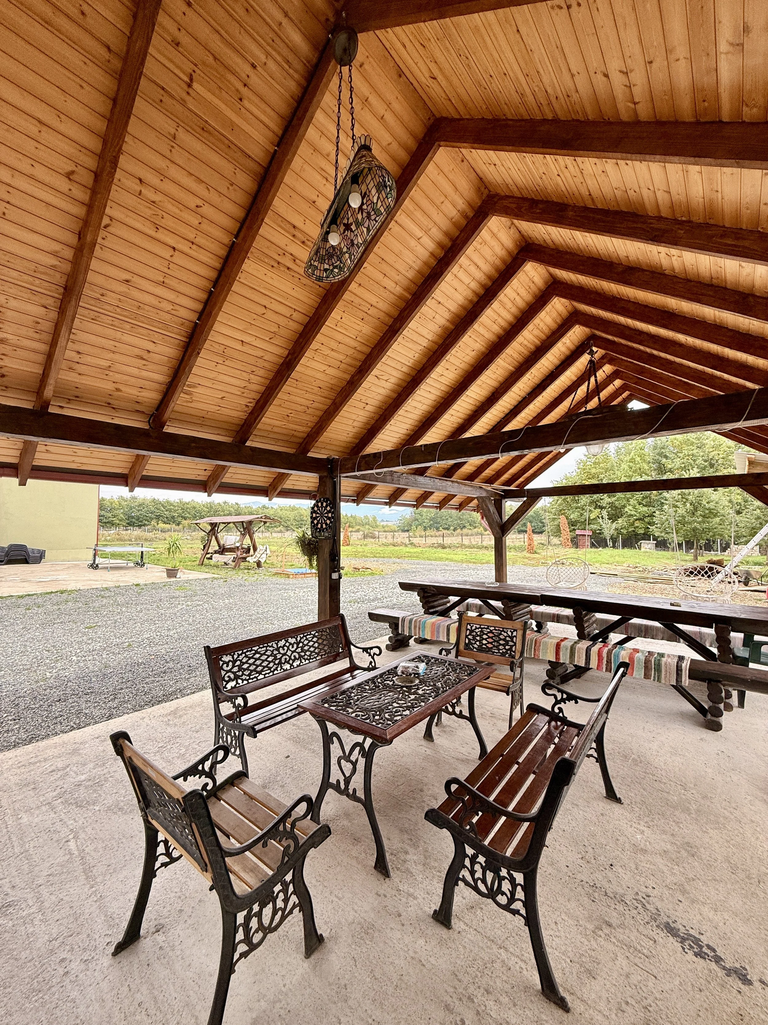 Outdoor covered patio with metal and wooden furniture, including chairs, benches, and tables, with a gravel yard and greenery in the background.