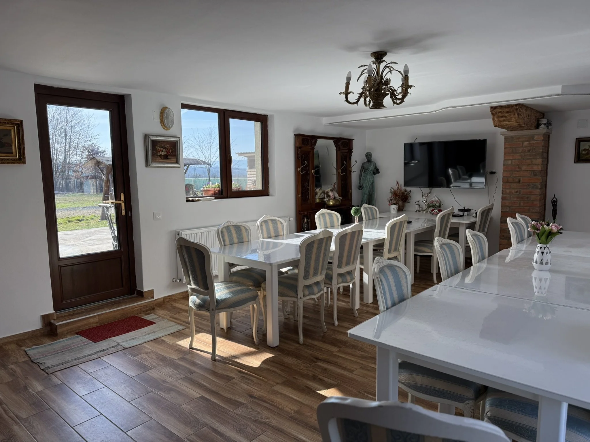 Bright dining room with white tables and striped upholstered chairs, large window and door showing outdoor scene, TV, chandelier, and decorative items.