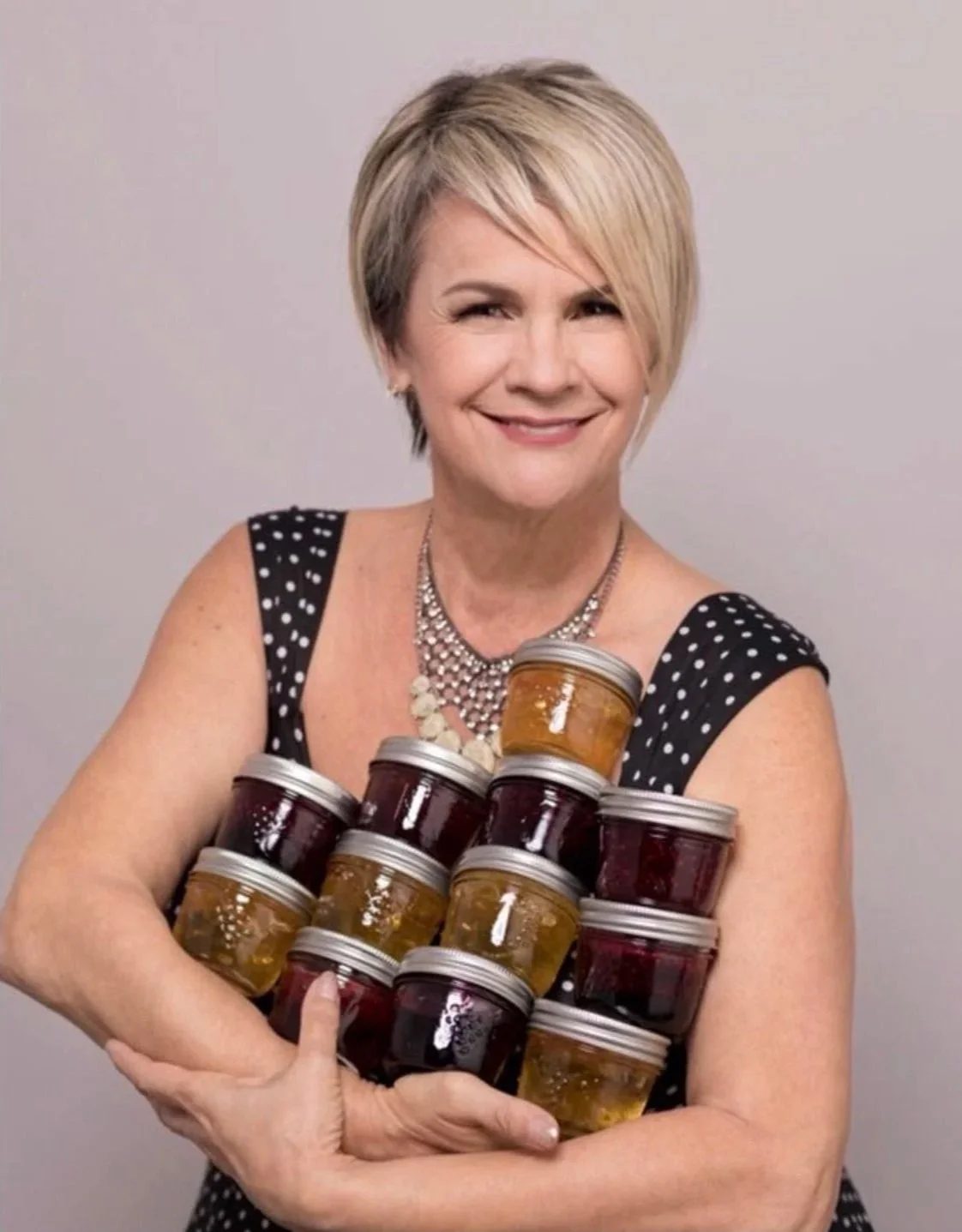 A smiling woman with short blonde hair in a black polka dot dress holding a pyramid of small jars of jam.