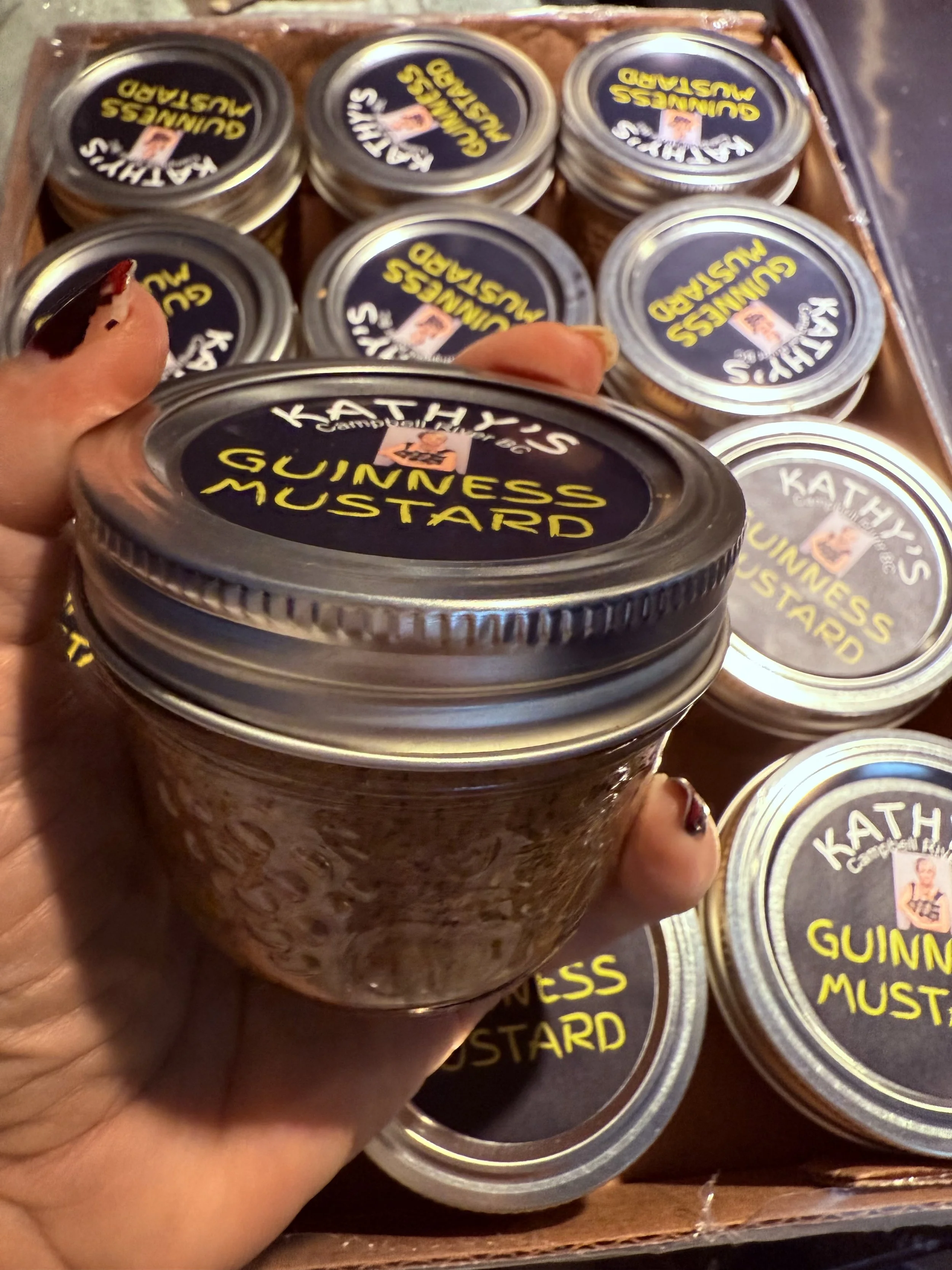A hand holding a jar of Kathy's Guinness Mustard, with more jars of the same mustard in a box behind it.