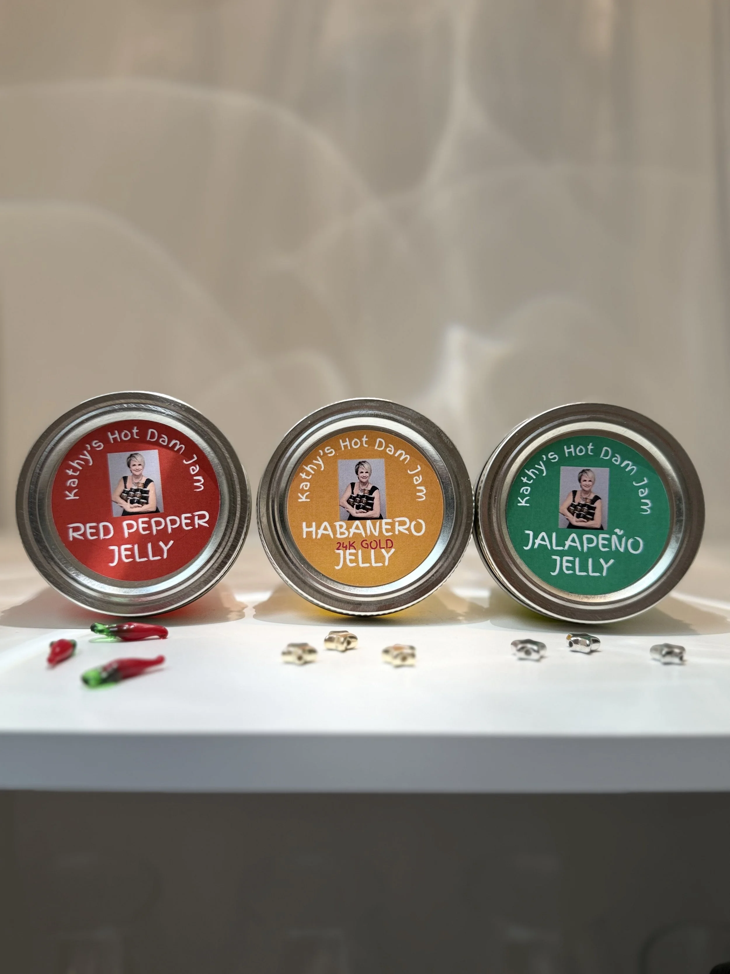 Three containers of jelly labeled as Kathy's Hot Damn well, with flavors Red Pepper Jelly, Habanero Jelly, and Jalapeño Jelly. Small decorative items resembling chili peppers and bones are placed in front of the containers.