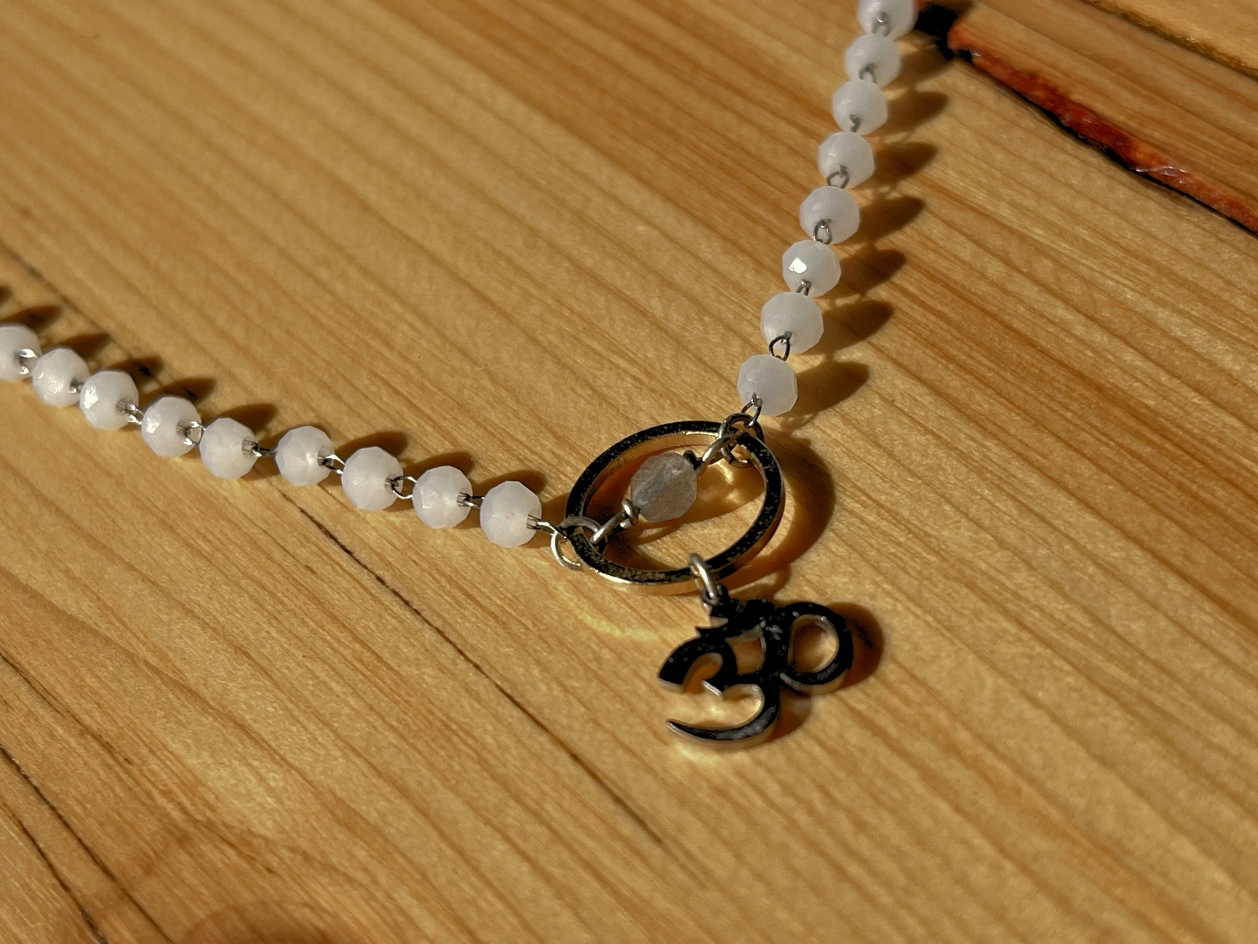 A beaded necklace with small white beads, a small grey stone, a metal ring, and a black 'Om' pendant lying on a wooden surface.