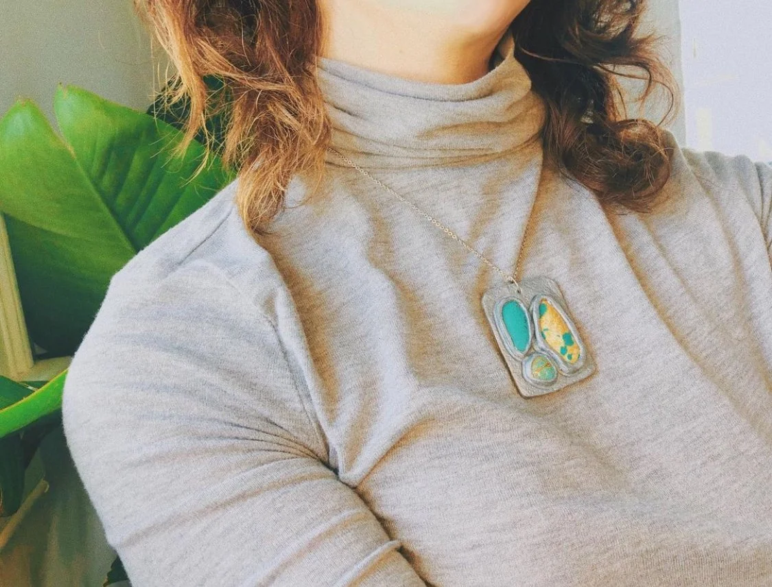 Close-up of a woman wearing a gray turtleneck shirt and a pendant necklace with colorful abstract shapes, near a large green plant.