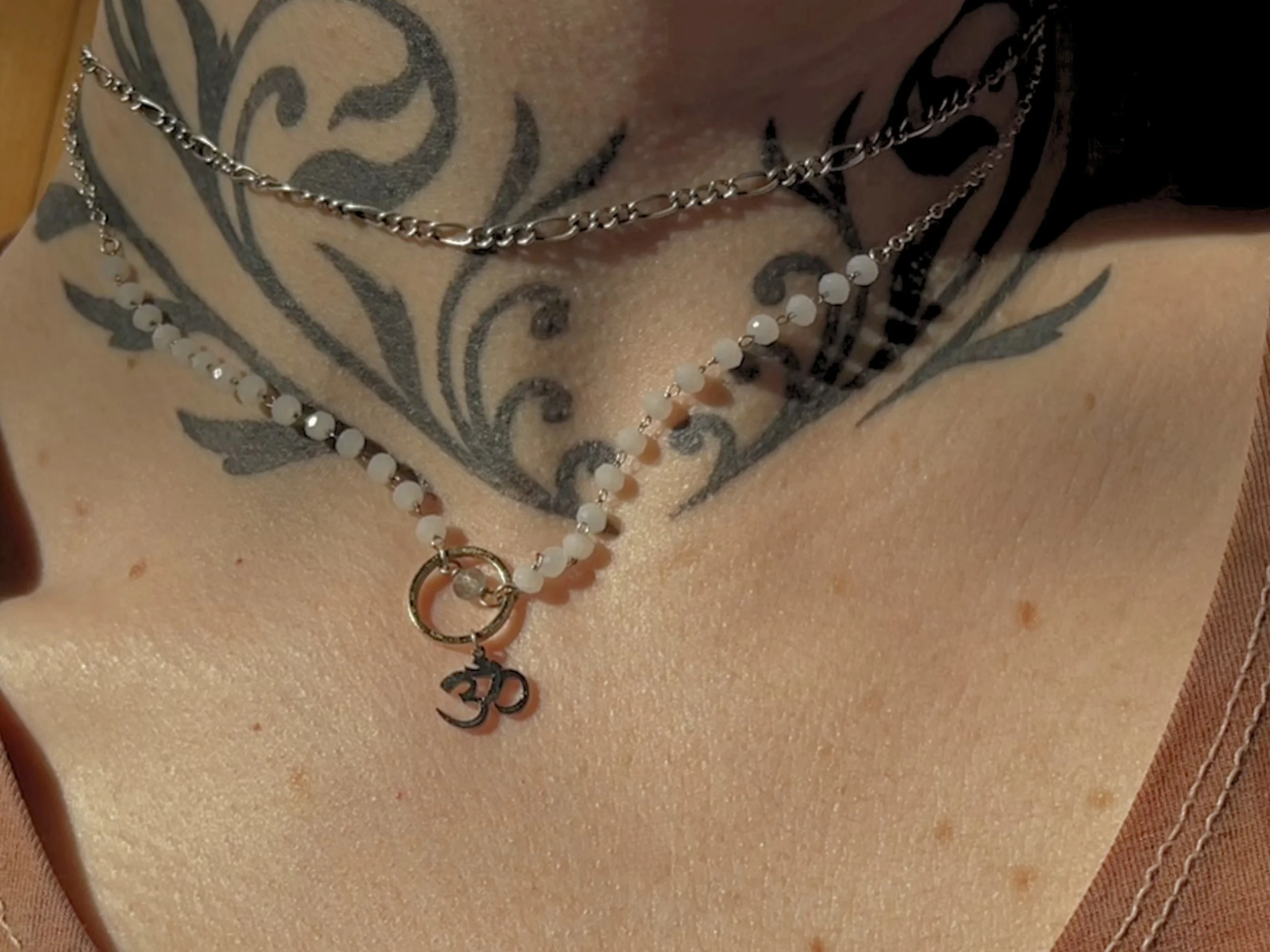 Close-up of a person's neck with two necklaces, one with white beads and a pendant with the 'Om' symbol, and the other a silver chain, against a person's chest with a large black tattoo.