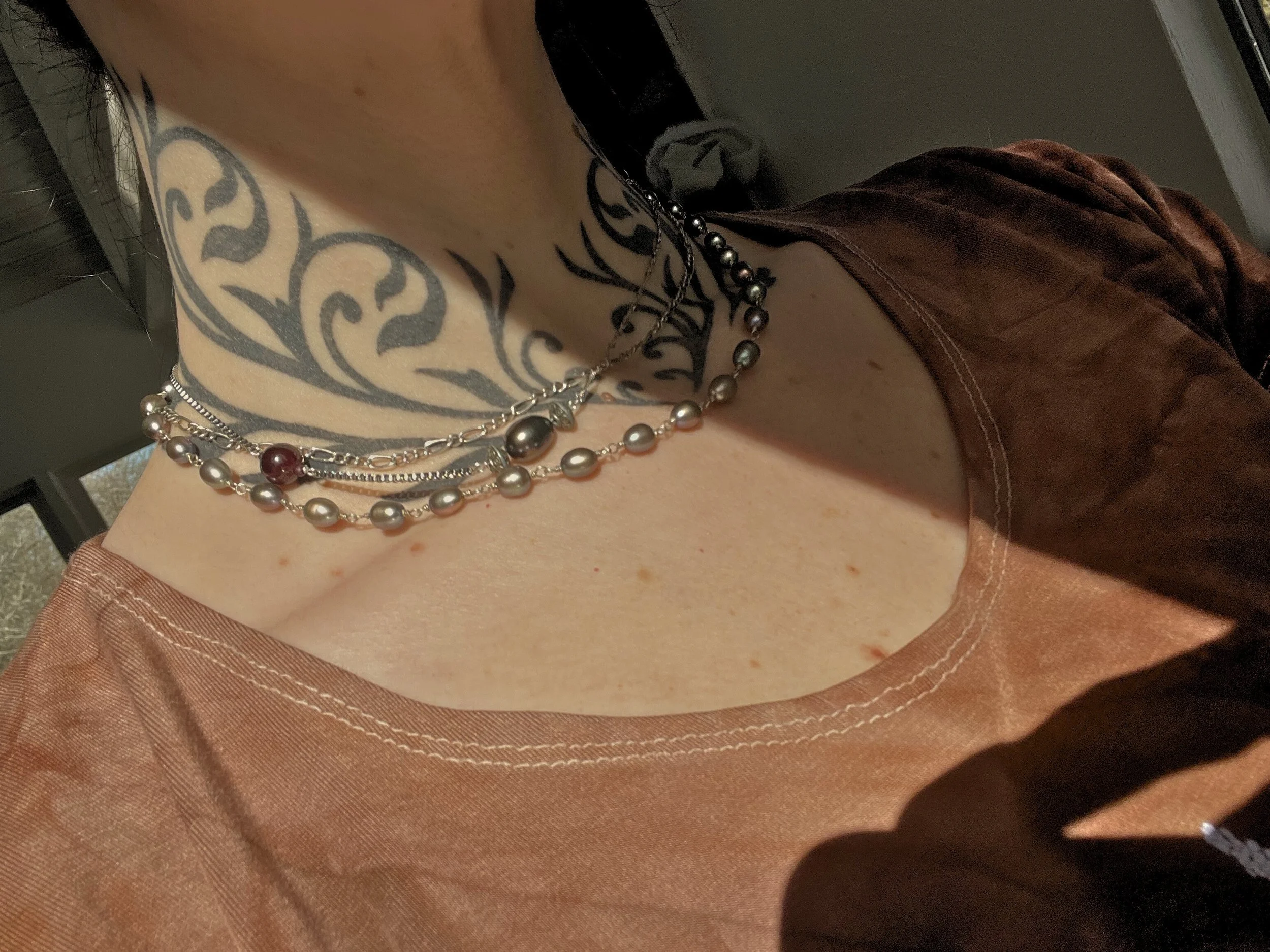 Person wearing multiple layered pearl and silver necklaces, a large tattoo on their chest, and a brown shirt.