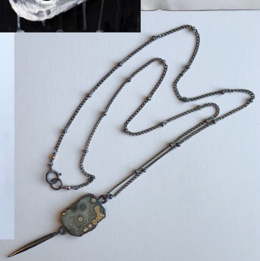 A necklace with a pendant shaped like a wall clock and a long, thin, pointed tail on a white surface.