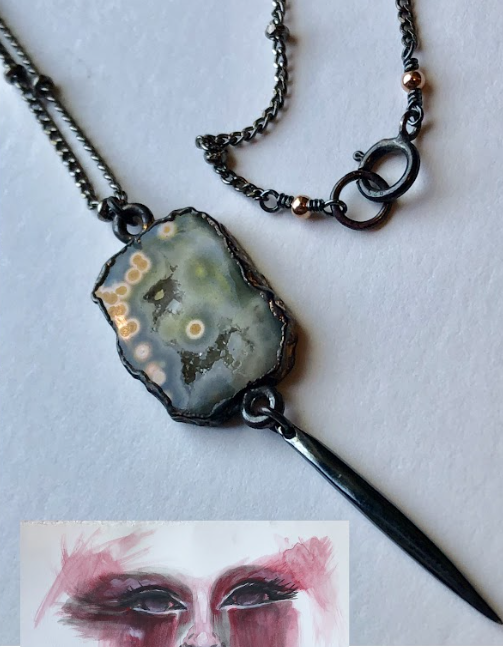 A necklace with a polished stone pendant and a metal needle attached, displayed on a white surface. A watercolor portrait of a face with dark makeup is partially visible below the necklace.