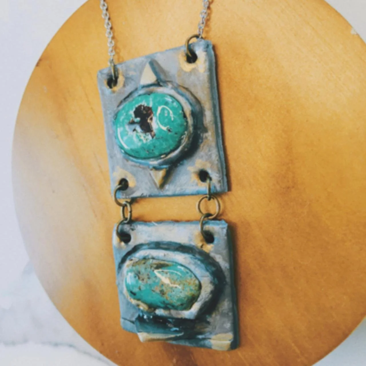Two vintage metal tiles with turquoise glass embellishments, connected by metal rings, hanging against a light brown wooden background.