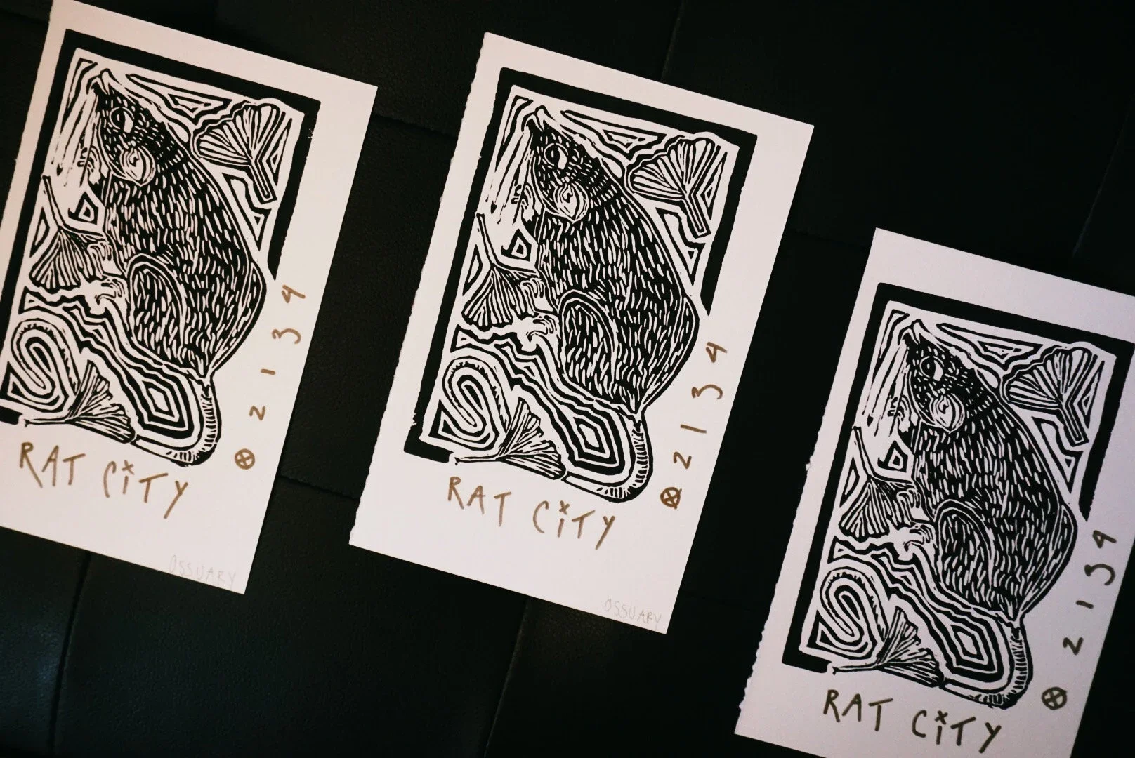 Three black and white prints on paper featuring stylized rat illustrations with the words 'RAT CITY' and '2134' written in brown, laid out on a black surface.