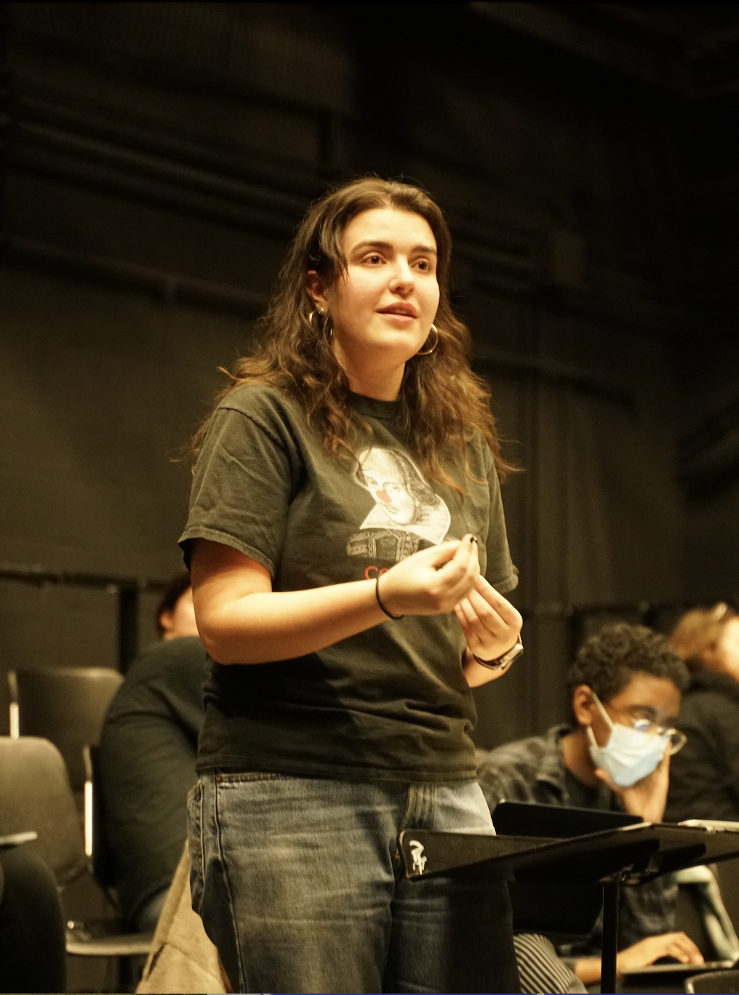 Young woman standing and speaking in a dimly lit room with seated audience, one wearing a face mask.