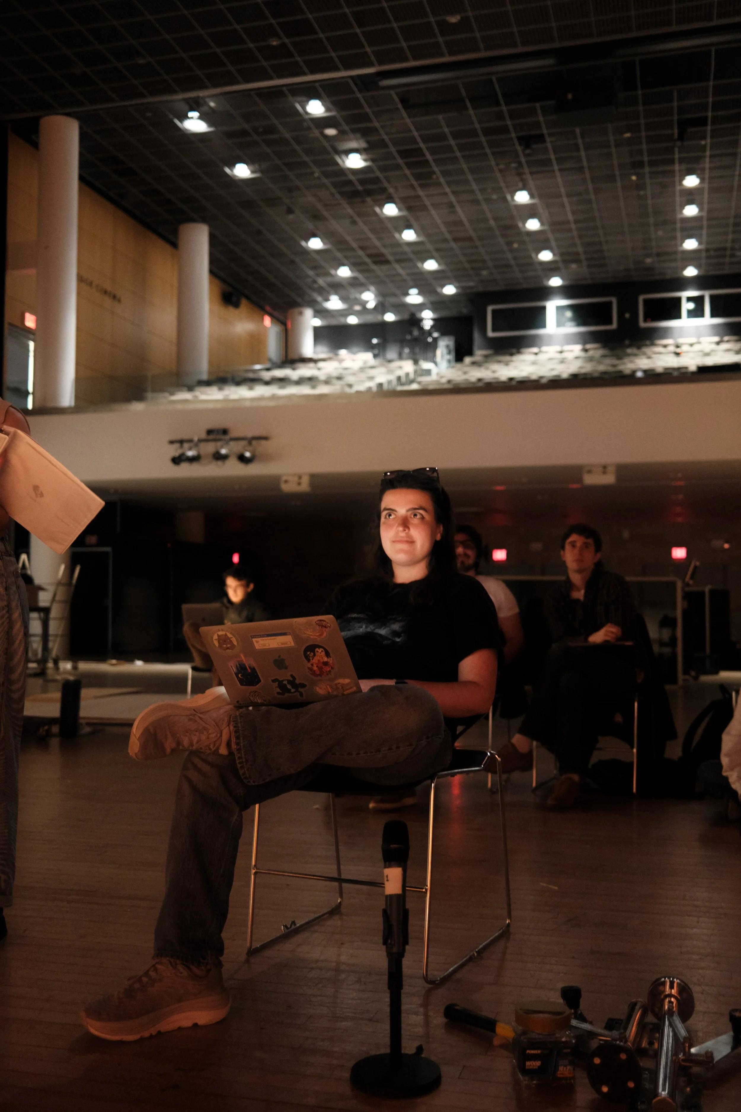 A young woman sitting on a chair in a dimly lit auditorium or lecture hall, with a laptop on her lap that is decorated with stickers, and surrounded by other seated people.