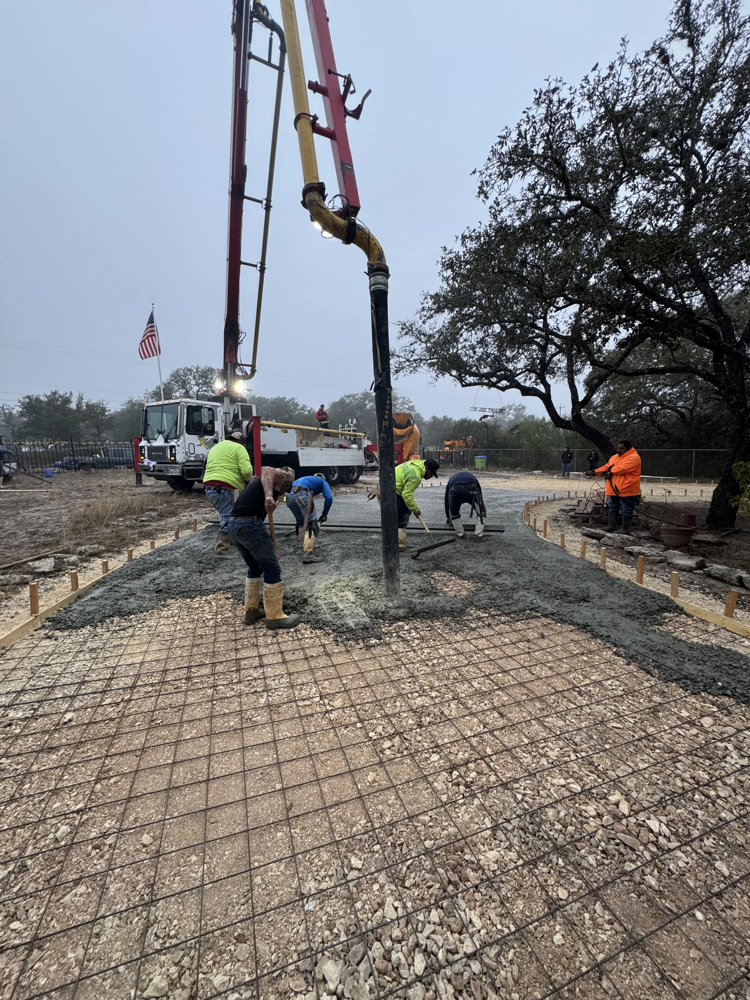 Construction workers pouring concrete on a building site, with a concrete pump truck and a tree in the background.